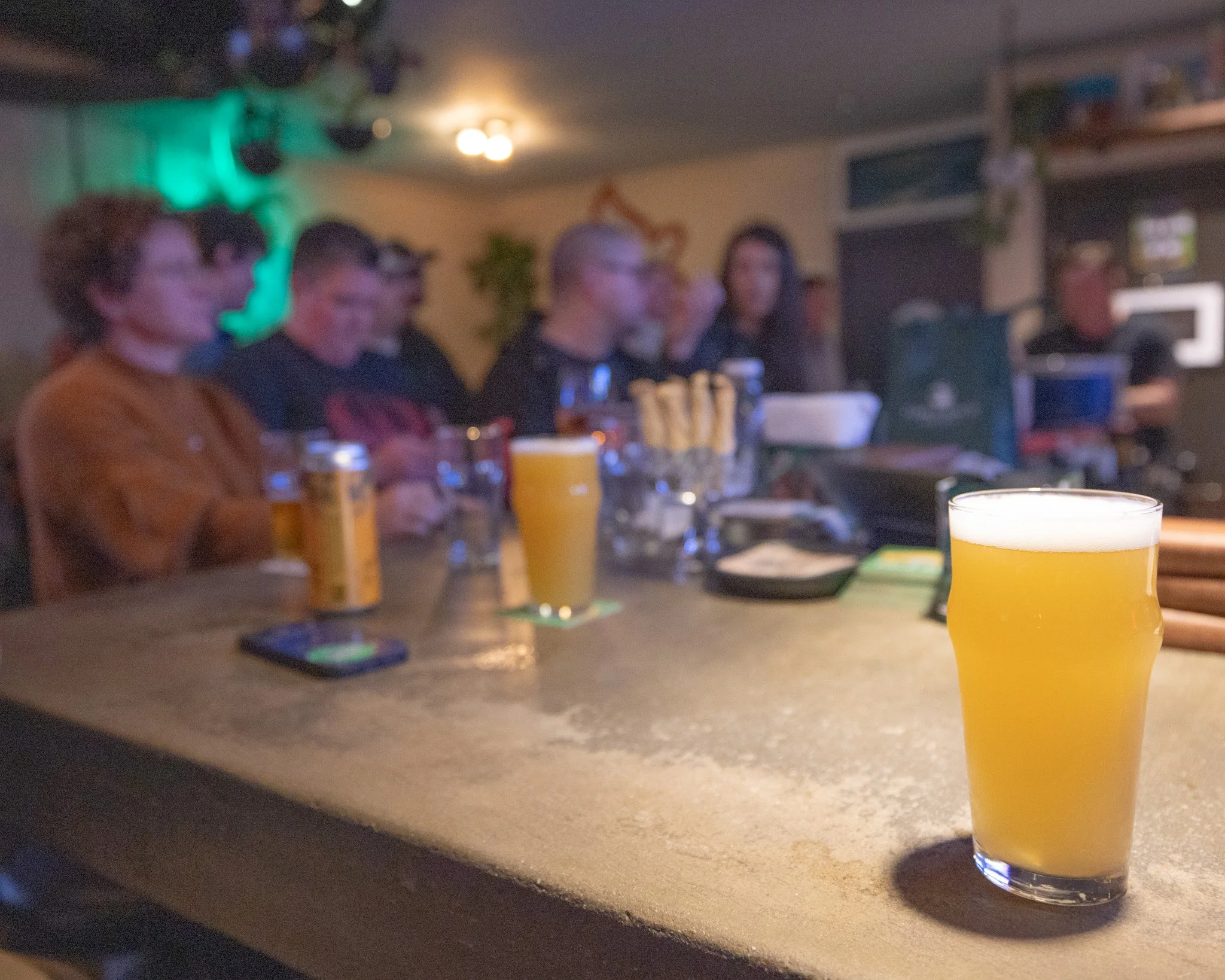 A glass of yellow beverage on a bar or countertop with other drinks and snacks in the background, in a dimly lit setting.