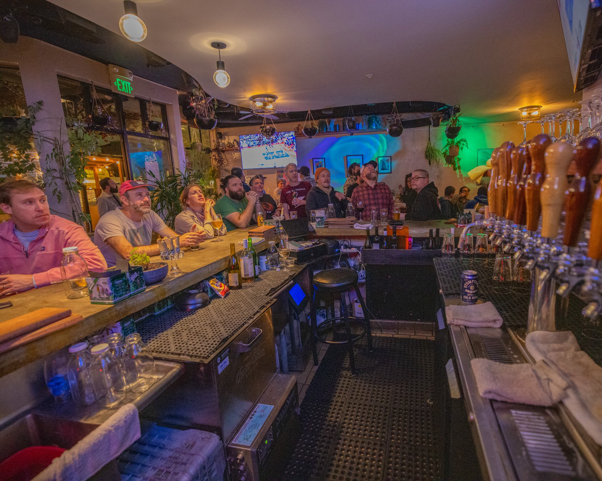 A lively bar or pub with a group of people seated at the bar and others standing, watching a large flat-screen TV mounted on the wall displaying a sporting event. The bar has beer taps with multiple handles, glasses, and bottles. The room is decorate