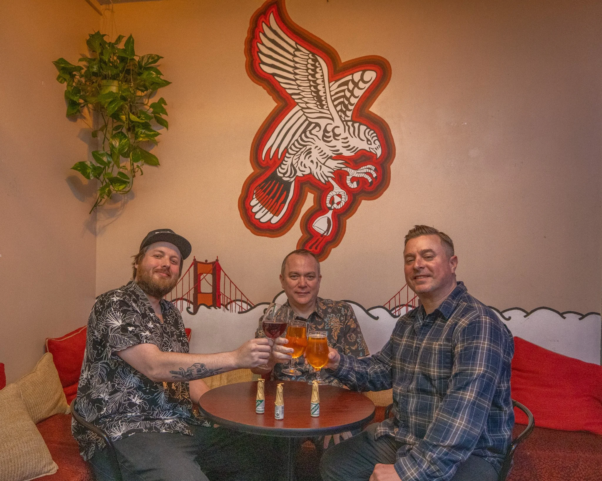 Three men are smiling and holding drinks in a cozy room with a table, red cushions, and a large poster of an eagle in front of the Golden Gate Bridge on the wall. There is a plant hanging in the corner.