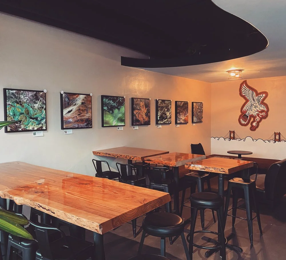 Interior of a cafe or restaurant with wooden tables, black chairs, and colorful abstract artwork on the wall. A mural of a bridge and an eagle is painted on the wall.