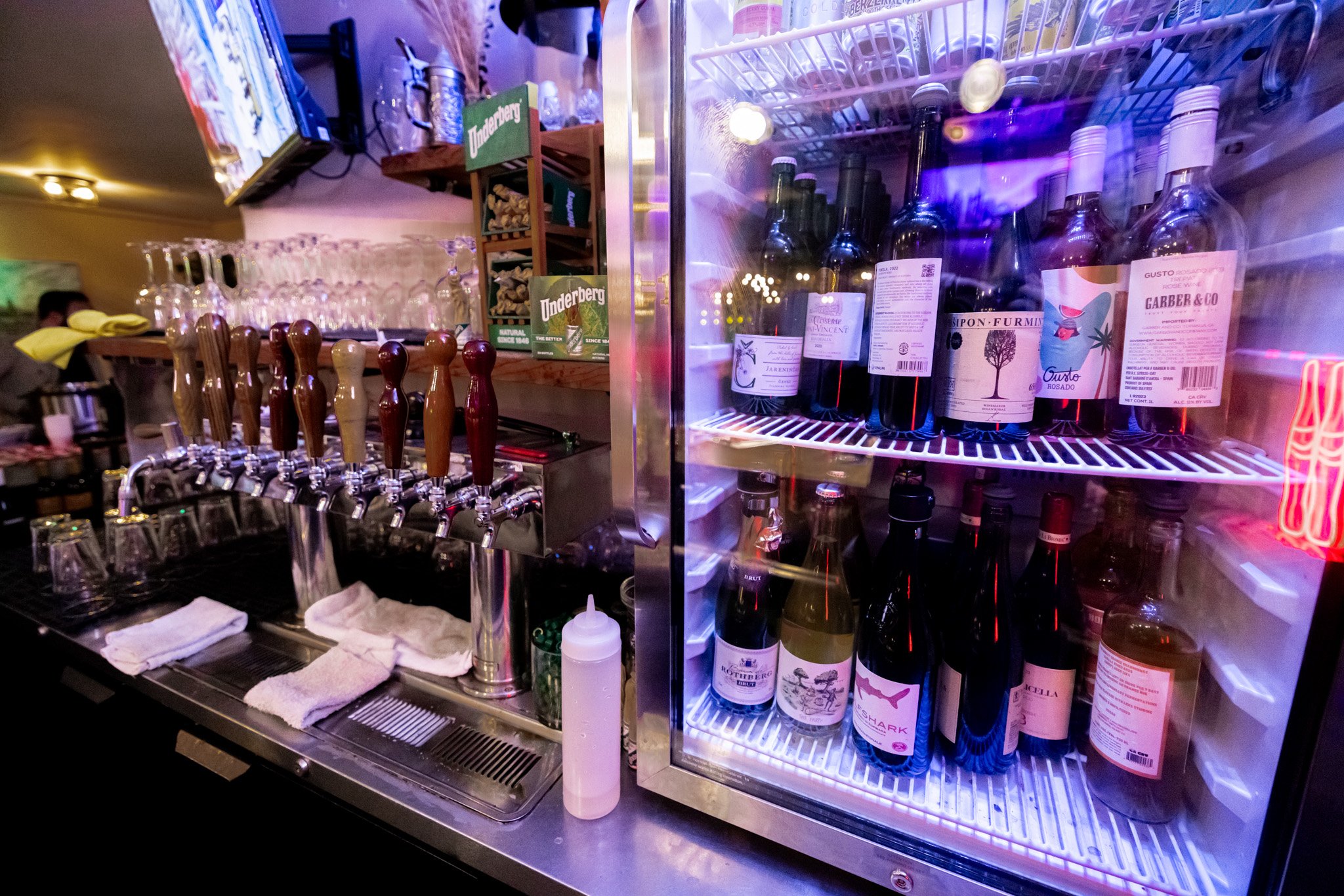 A bar counter with beer taps, glasses, and a fridge with wine bottles inside