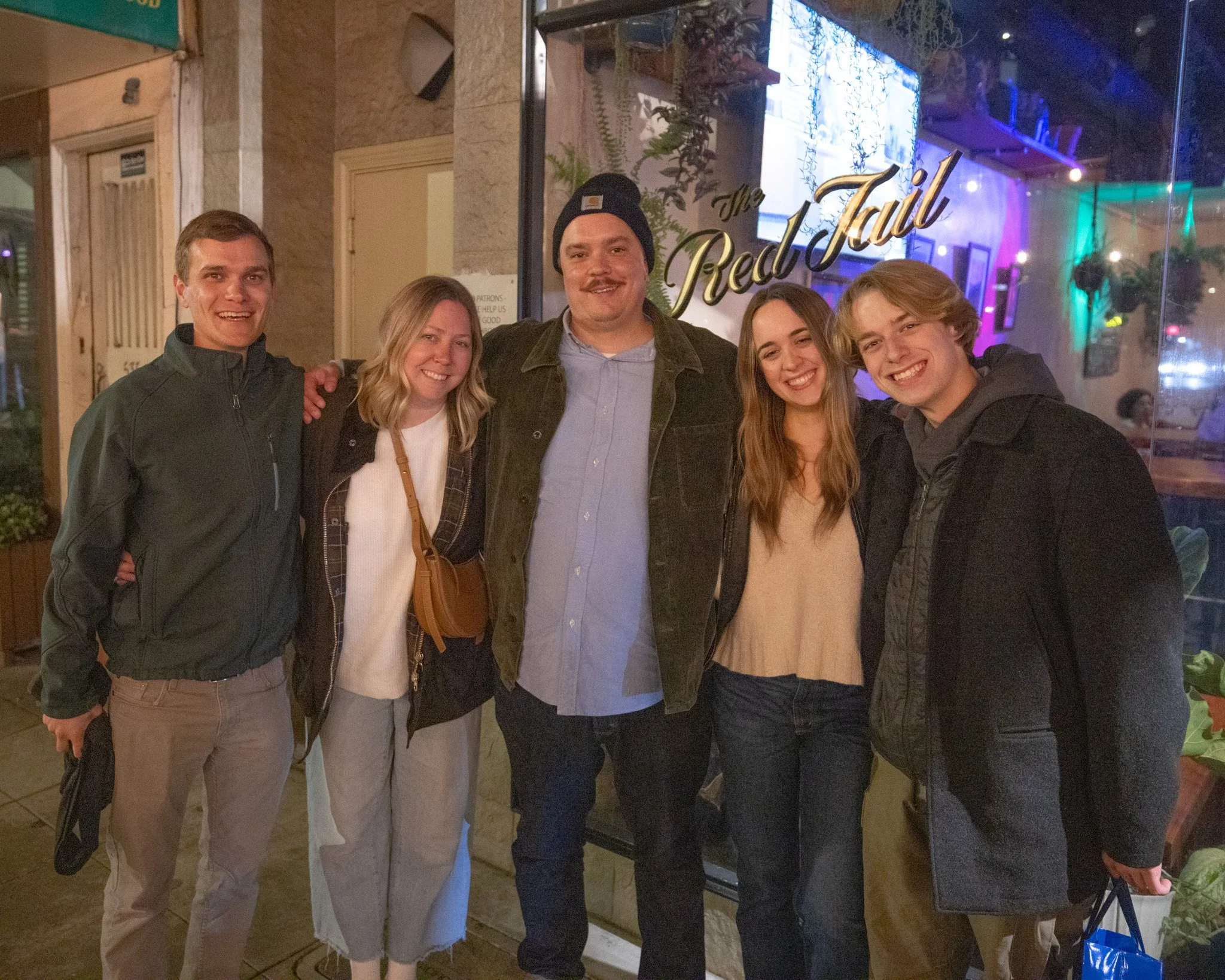 Five people are standing together inside a restaurant, smiling at the camera. The restaurant has a decorated glass window with the words 'The RedTail' written on it, and purple and green lights in the background.