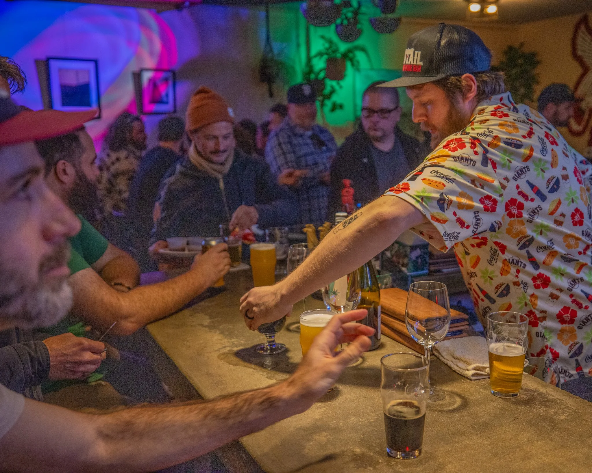 Group of people socializing in a bar with drinks on the counter, colorful lighting, and wall decorations.
