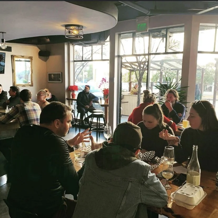 People dining and socializing inside a cozy restaurant with large windows, natural light, and potted plants.