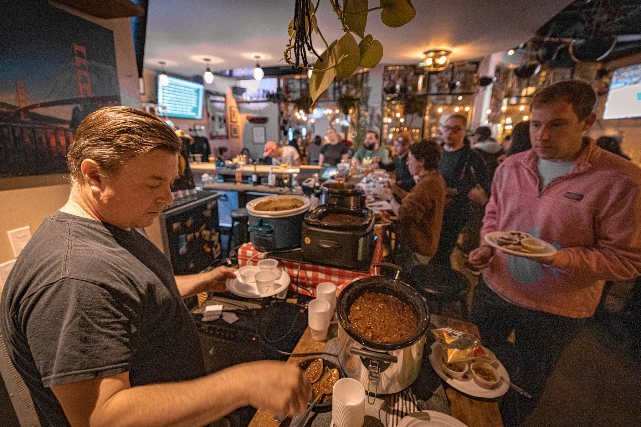A lively indoor gathering at a restaurant with people serving themselves food from a buffet table. The scene includes a man in a gray shirt holding a plate and tongs, a man in a pink jacket holding a plate, and several other people dining and sociali