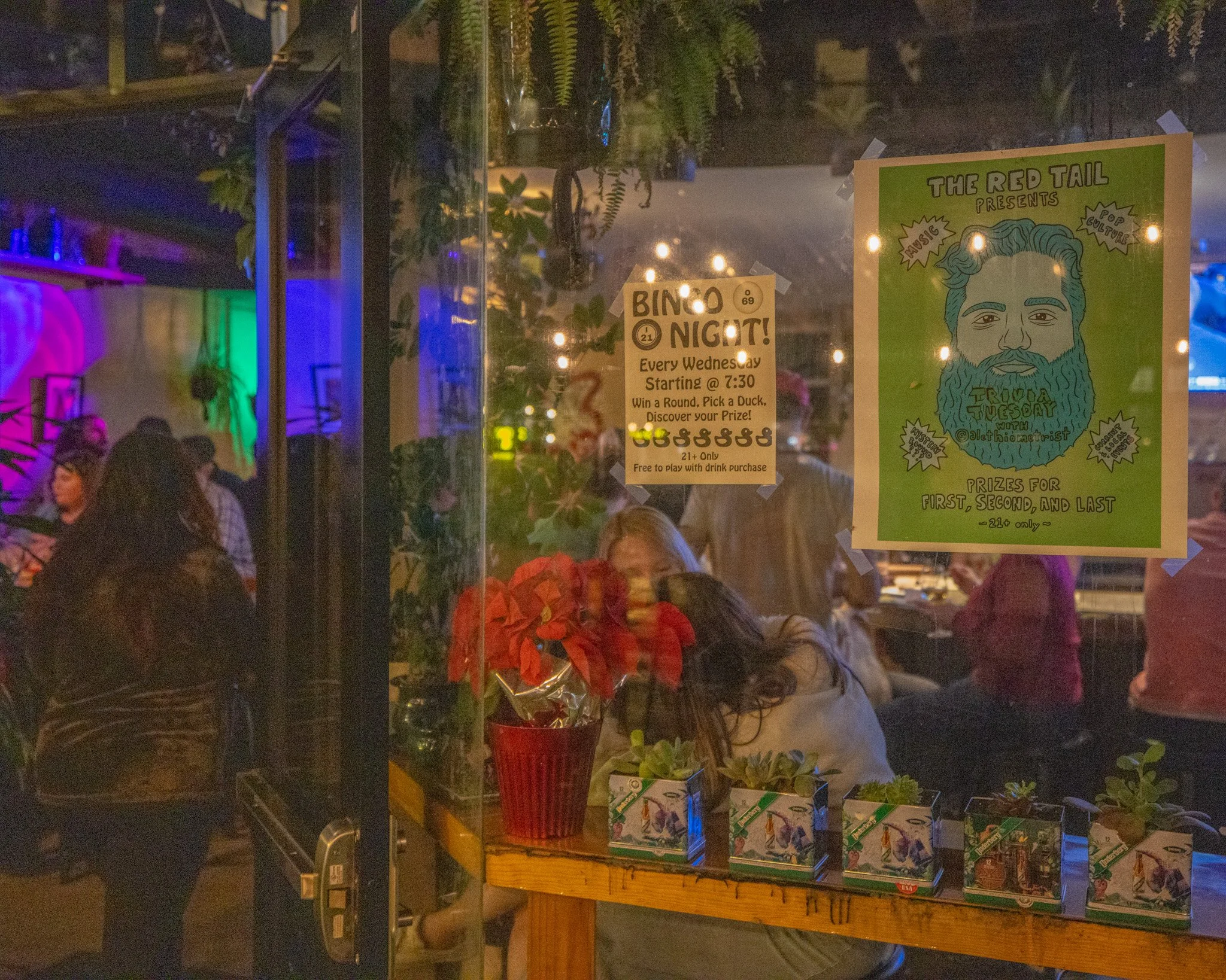 Interior view of a retail store or event space with colorful neon and string lights, showcasing various products including plants, garden supplies, and promotional posters. There are people browsing inside.