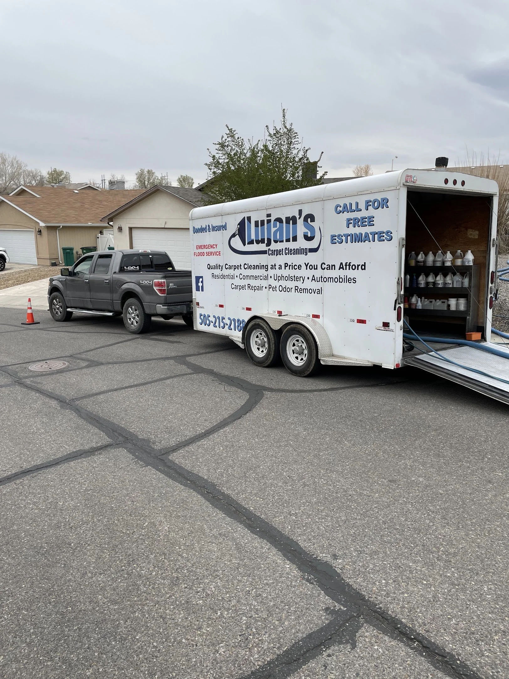 A black pickup truck parked next to a white mobile carpet cleaning trailer on a residential street. The trailer has advertising for Lujan's Carpet Cleaning, including services like carpet cleaning, pet odor removal, and estimates. Houses with garages are visible in the background under a cloudy sky.