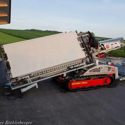 A damaged tracked industrial machine or equipment on a paved road in a rural area with grassy fields and a partly cloudy sky in the background.