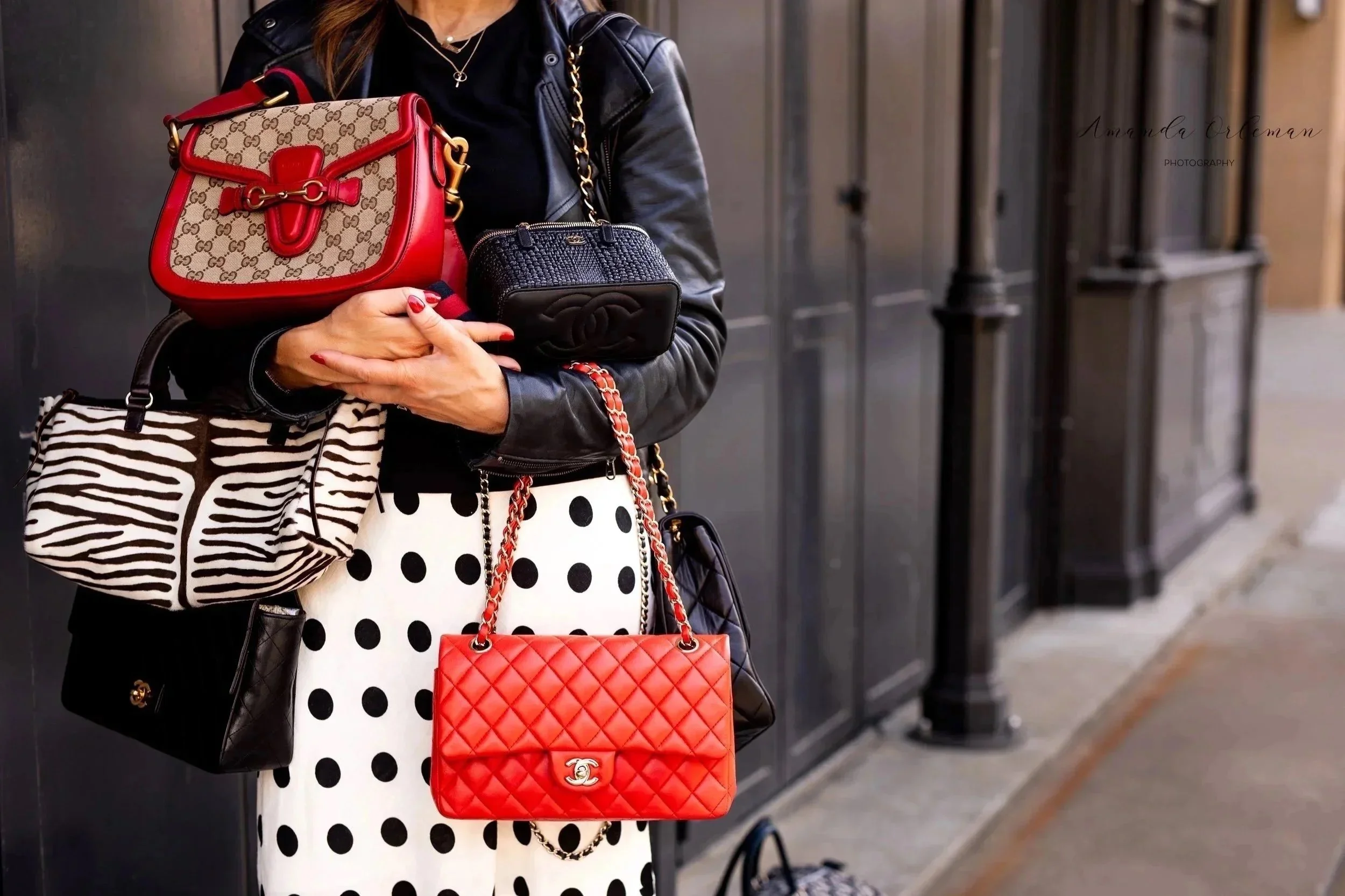 Close-up of a woman wearing a black leather jacket, holding multiple designer handbags, including a red and beige Gucci bag, a black Chanel bag, and a small red Chanel bag, against a dark background.
