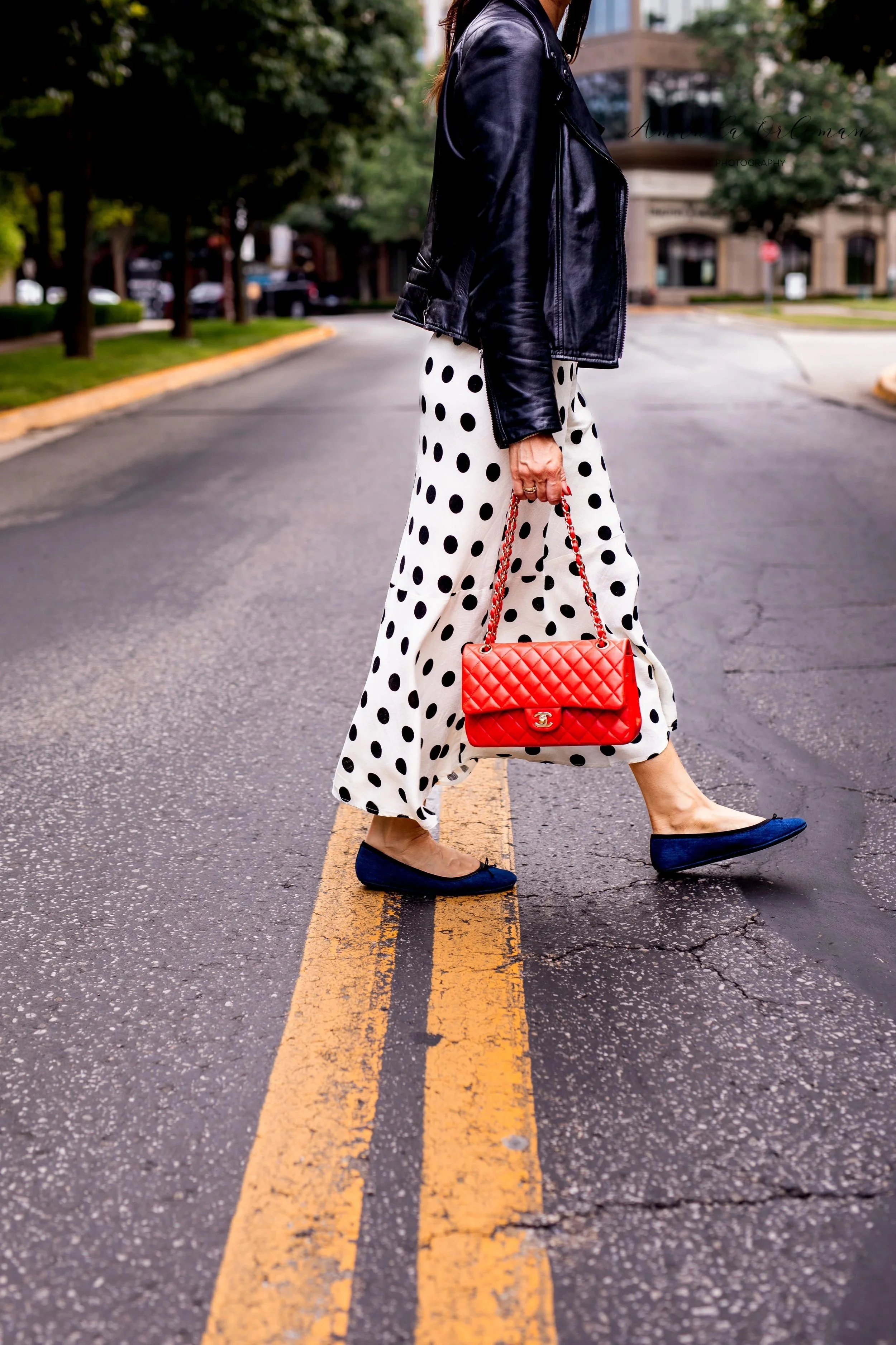 A woman walking across a street at a pedestrian crosswalk, holding a red quilted Chanel handbag, wearing a black leather jacket, polka dot skirt, and denim shoes.