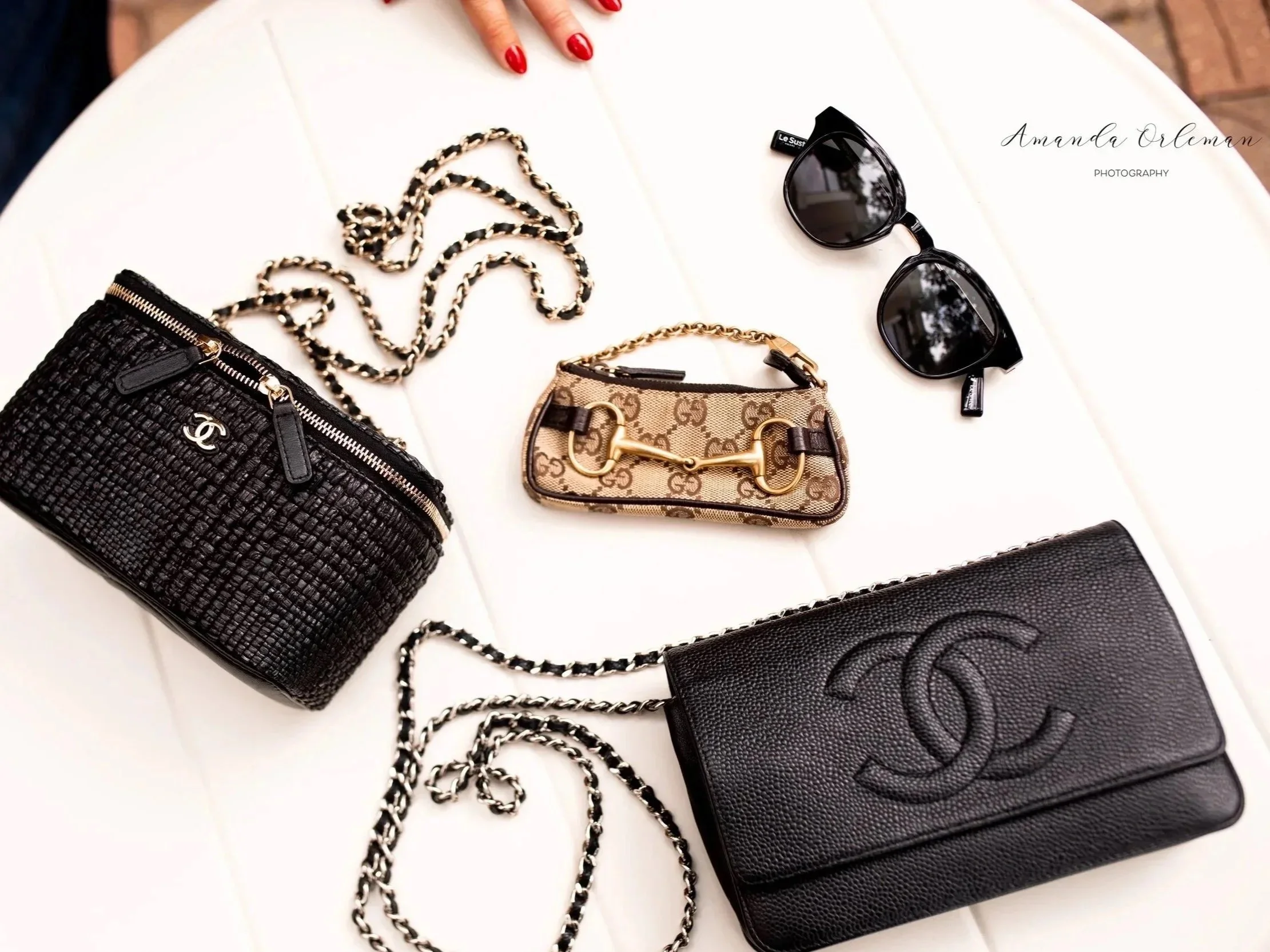 A flat lay of designer accessories on a white surface, including a black quilted Chanel wallet with gold zipper, a beige Gucci miniature handbag with gold hardware, a pair of black sunglasses, and some jewelry with black and gold chains. A person's hand with red nails is visible at the top of the image.