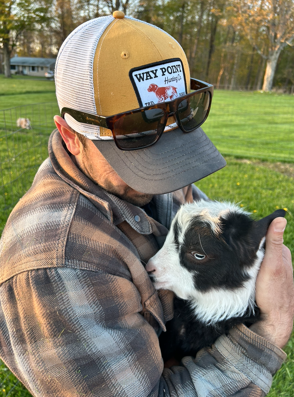 A man wearing a baseball cap with a patch logo, sunglasses, and a plaid jacket is holding and cuddling a black and white puppy outdoors on a grassy area with trees in the background.