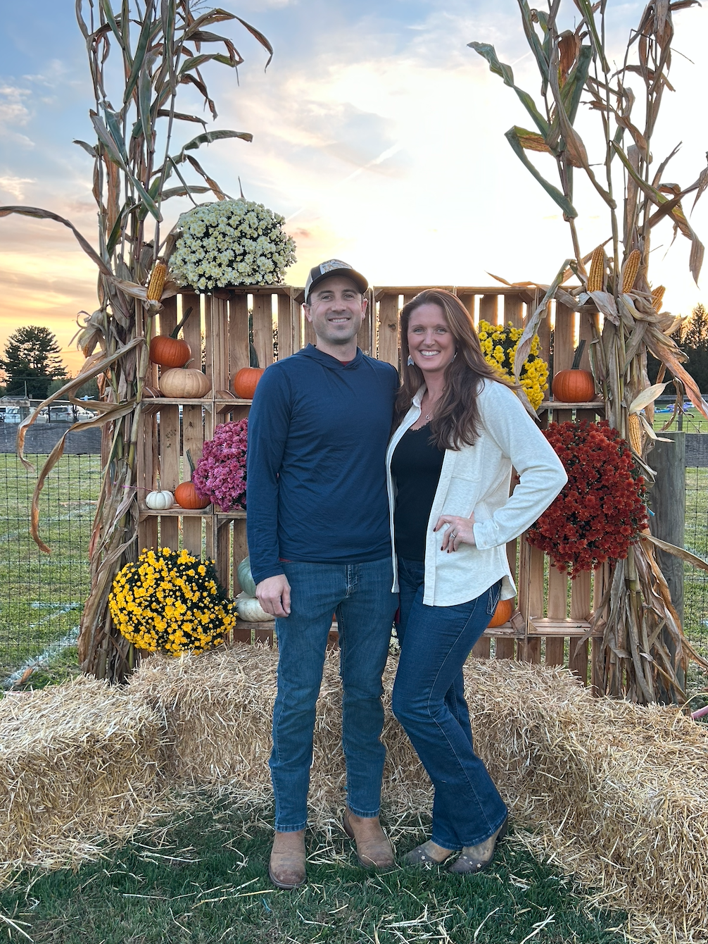 A smiling couple standing outdoors at an autumn festival or celebration, posing in front of a decorative wooden backdrop adorned with pumpkins, gourds, and colorful chrysanthemums. The scene is set at sunset with a partly cloudy sky.