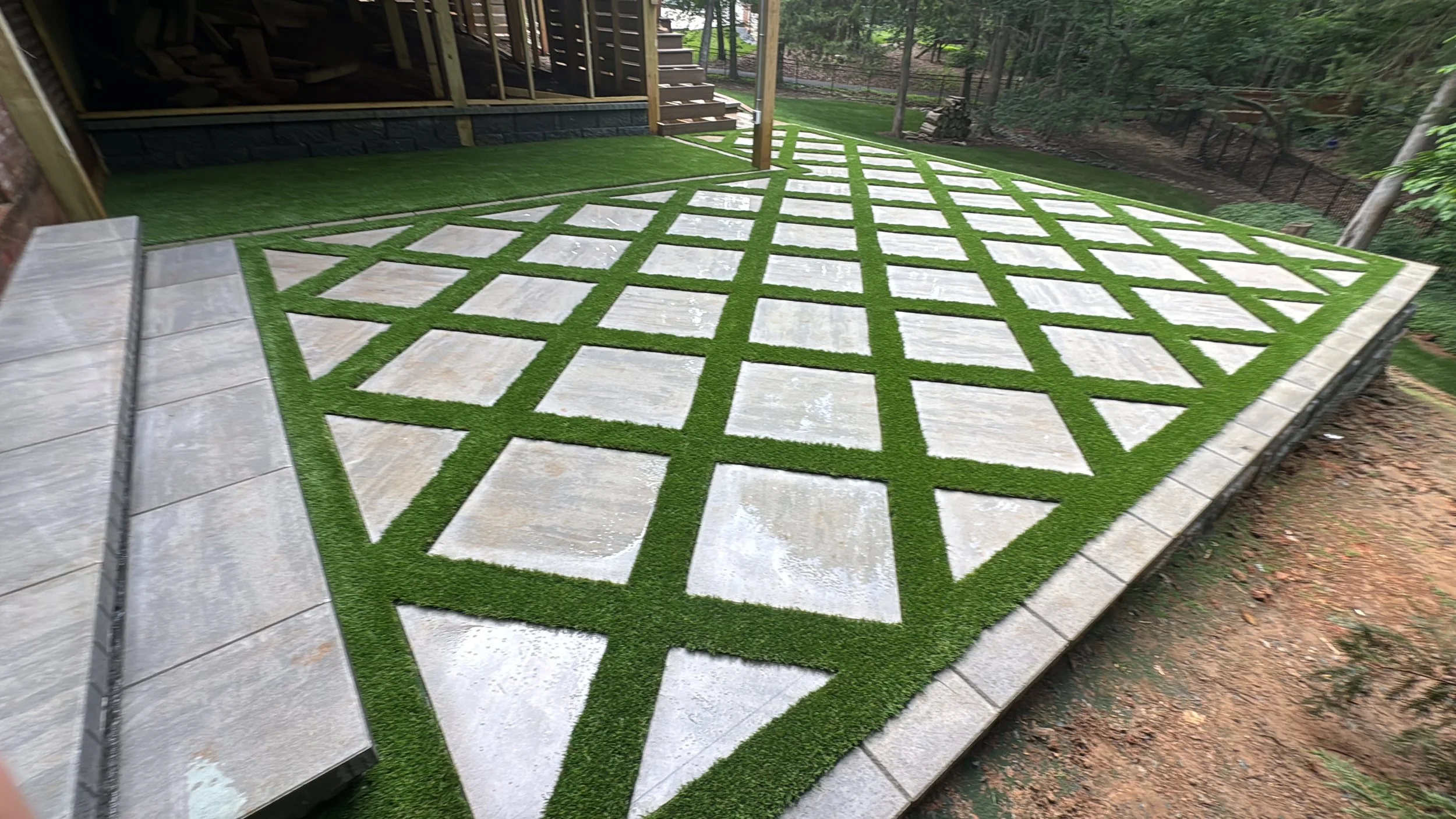 Newly installed concrete patio with green grass grid pattern in backyard, with stairs leading to an elevated deck and fenced wooded yard.
