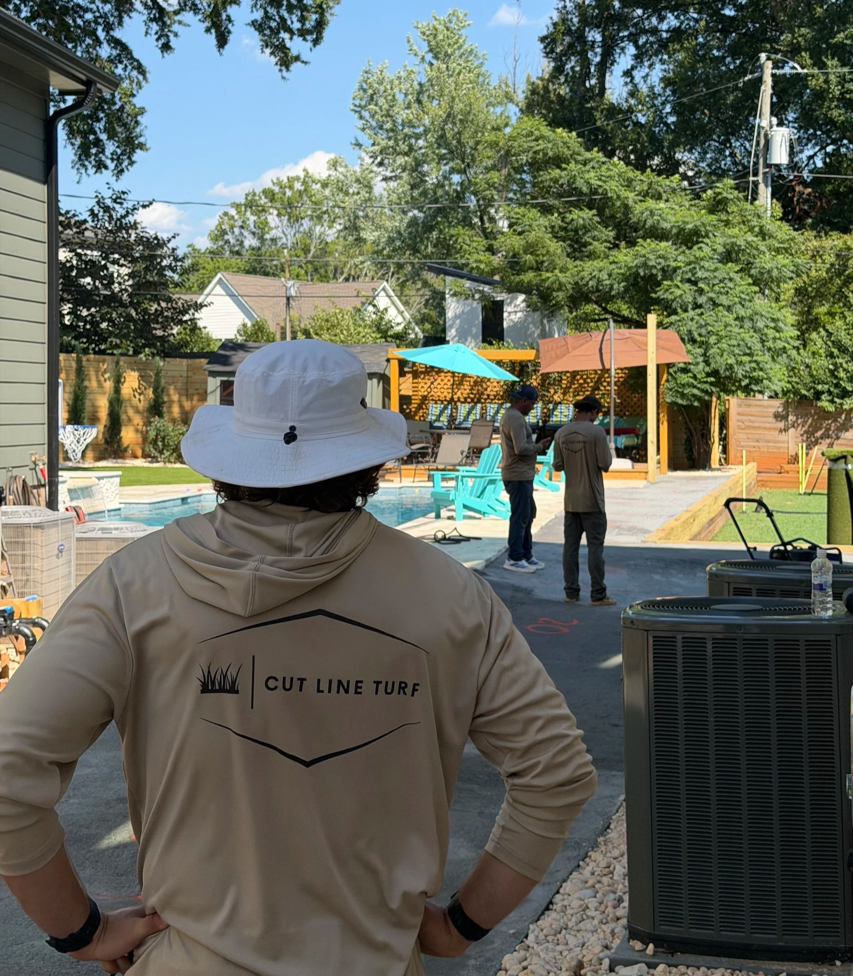Person wearing a beige jacket with a logo and the words "Cut Line Turf" on the back, standing outdoors near a fence, with a pool, patio furniture, and other people in the background.