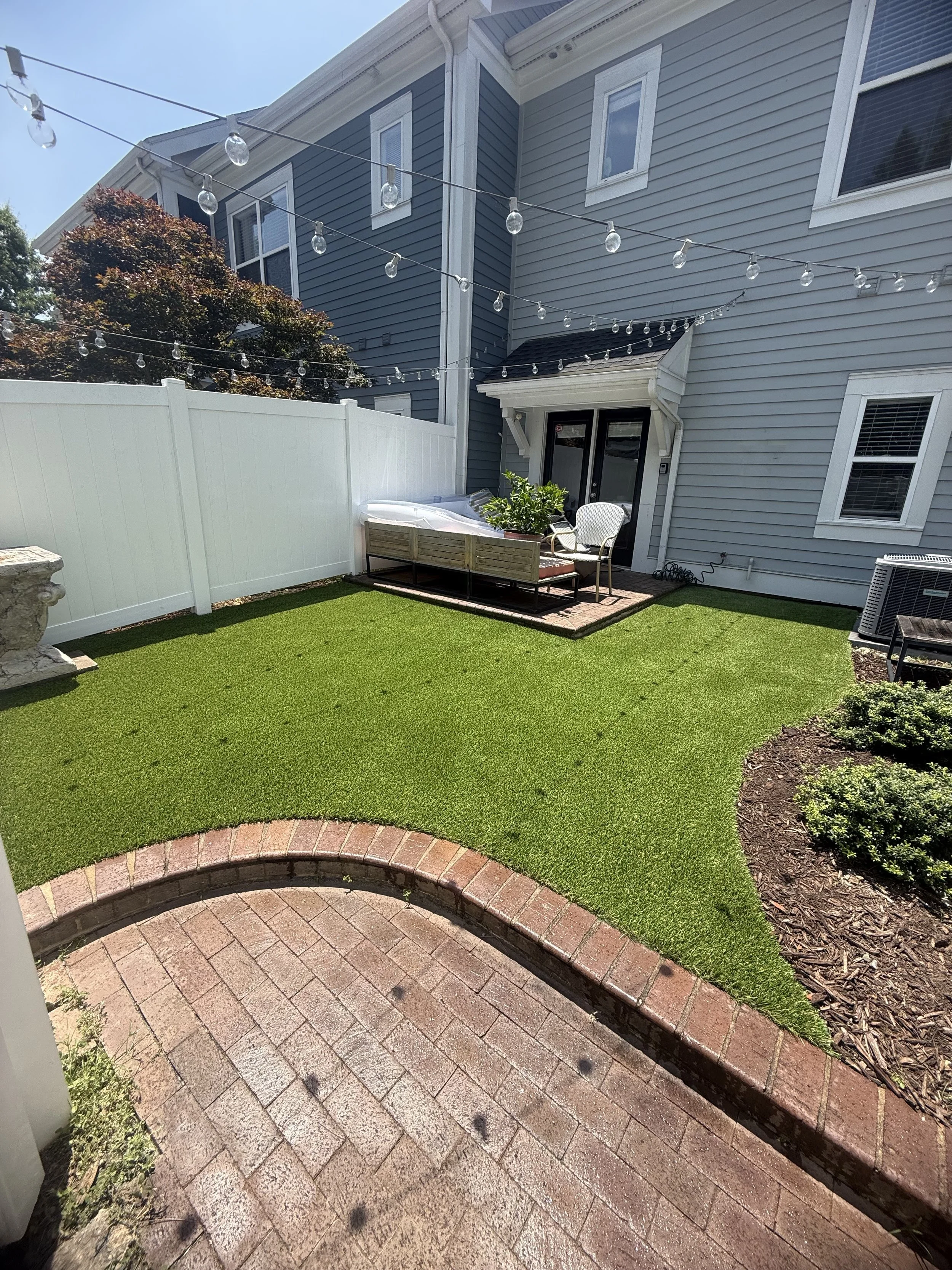 A backyard with a brick walkway, a grass lawn, a white fence, outdoor string lights, a potted plant, outdoor furniture, and a multi-story house with blue and gray siding.