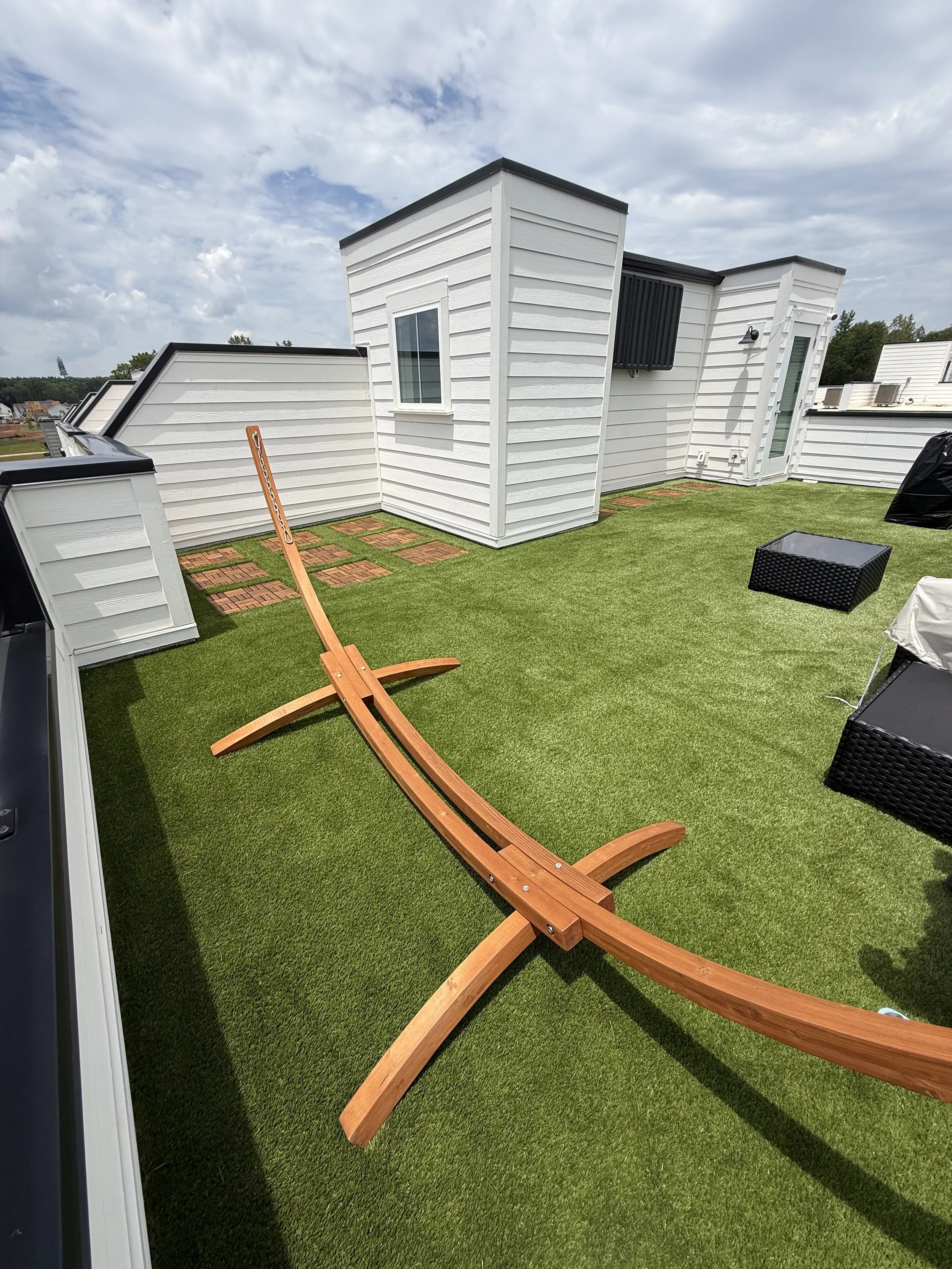 A rooftop deck with green artificial grass, a wooden lounge chair, black wicker furniture, and small brick pavers, surrounded by white siding buildings under a partly cloudy sky.