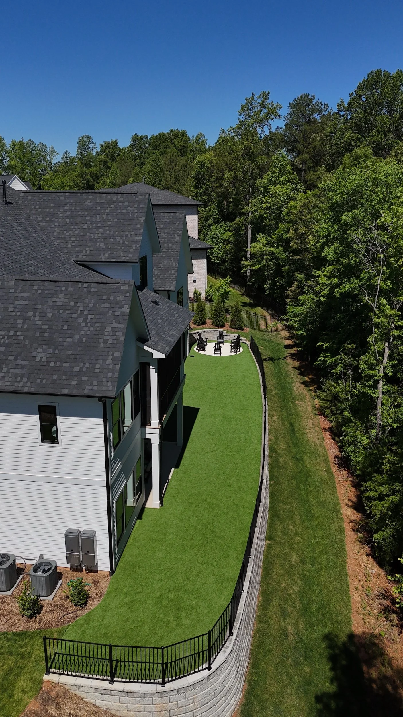 Aerial view of a modern house with a black roof, white siding, large windows, a backyard with a curved patio, outdoor seating, and a lush green lawn fenced with black metal, surrounded by trees and clear blue sky.