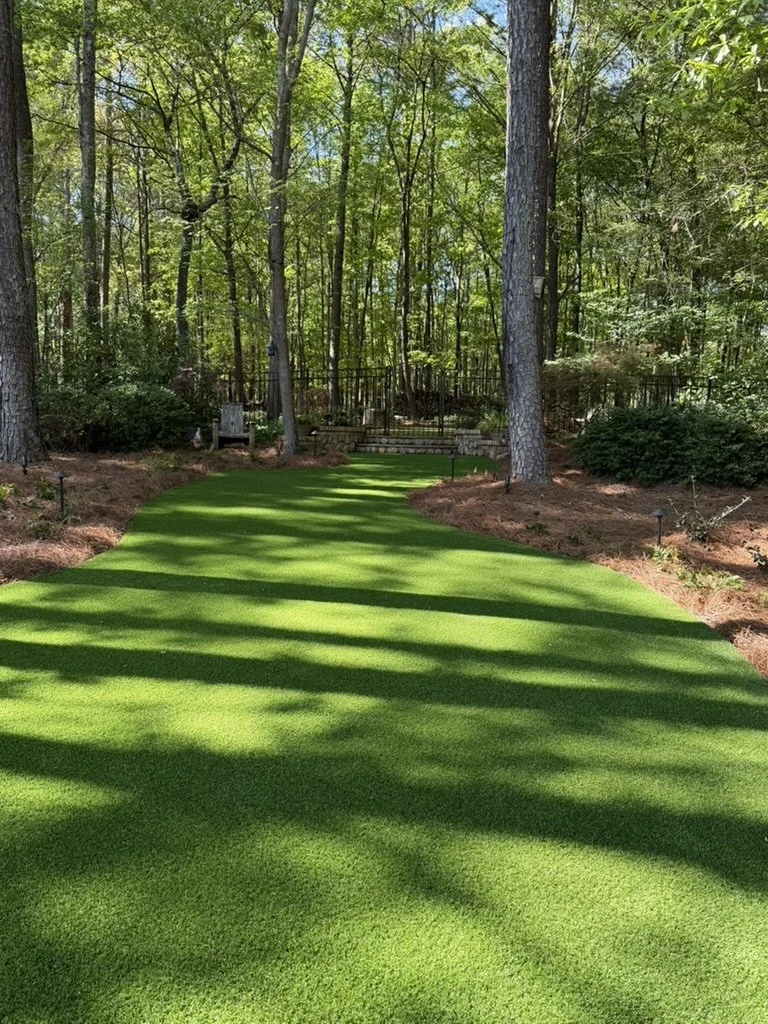 A backyard with a lush green artificial grass lawn surrounded by trees and bushes, with a wooden fence and garden chairs in the background.