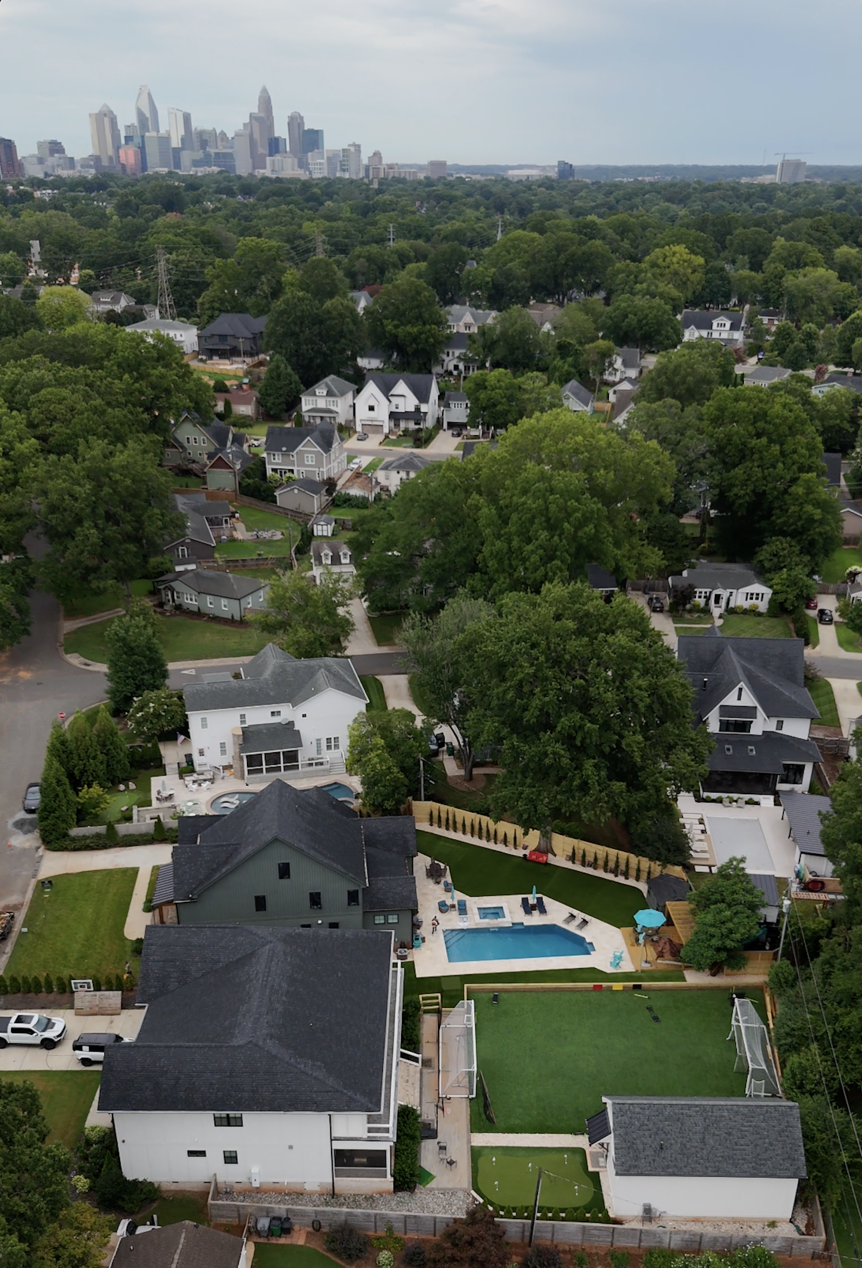 Aerial view of a suburban neighborhood with houses, a swimming pool, and green yards, with a city skyline in the distance under a cloudy sky.