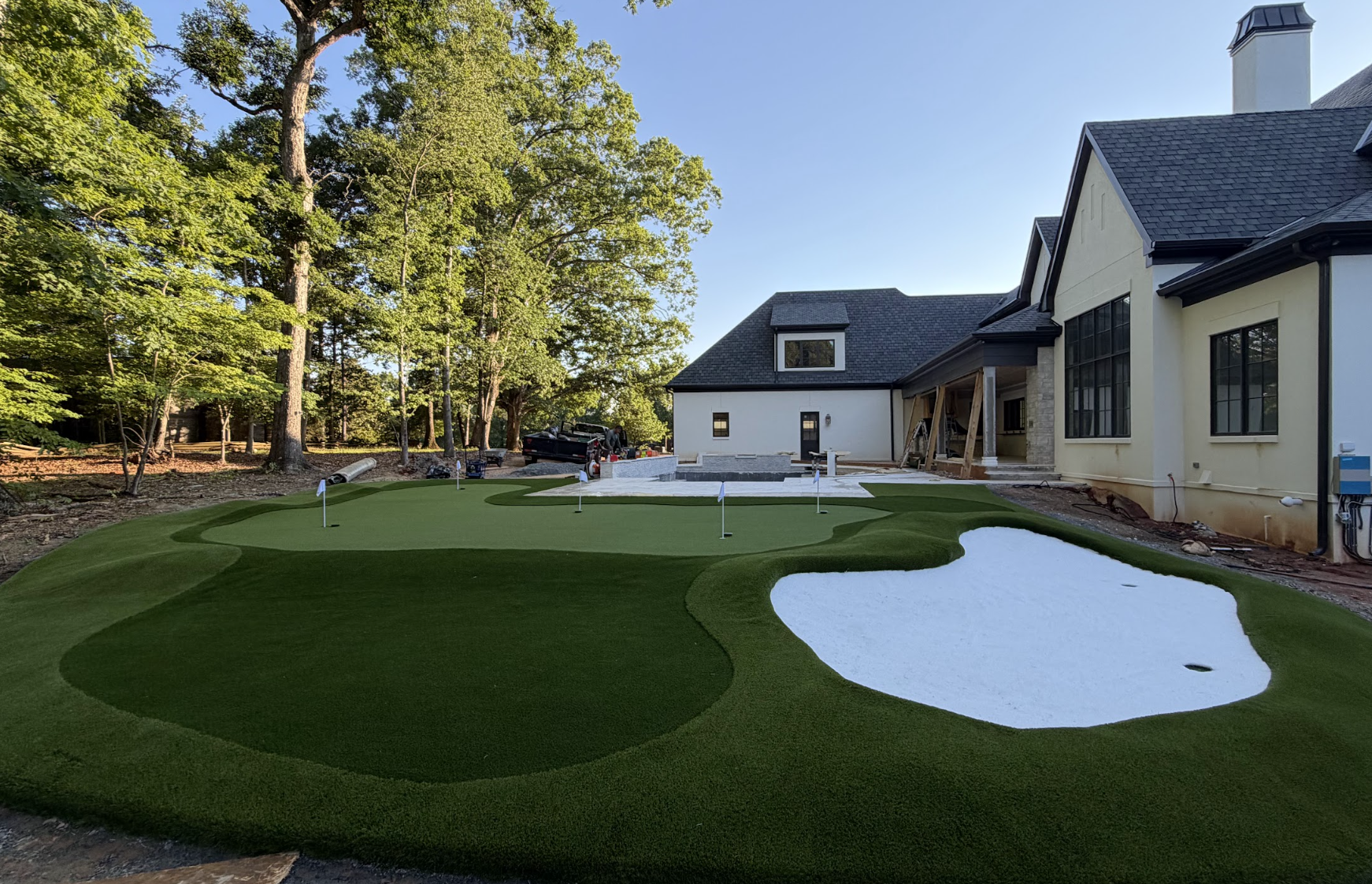 A backyard with a golf putting green and sand traps next to a house under construction, with construction tools and a truck visible