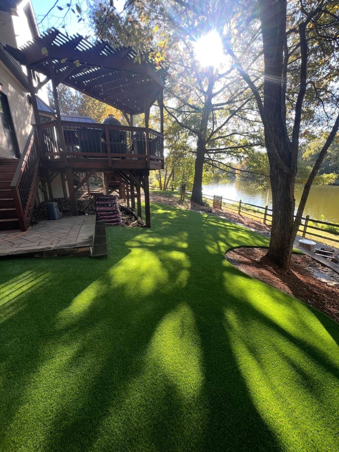 Backyard with bright green artificial grass, trees casting shadows, a deck with a grill, and a view of a river with a small dock.