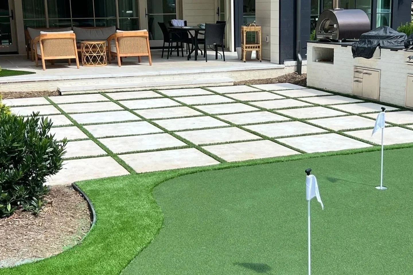 Backyard patio with outdoor furniture, a built-in grill, and a small putting green with flags, surrounded by landscaped grass and plants.