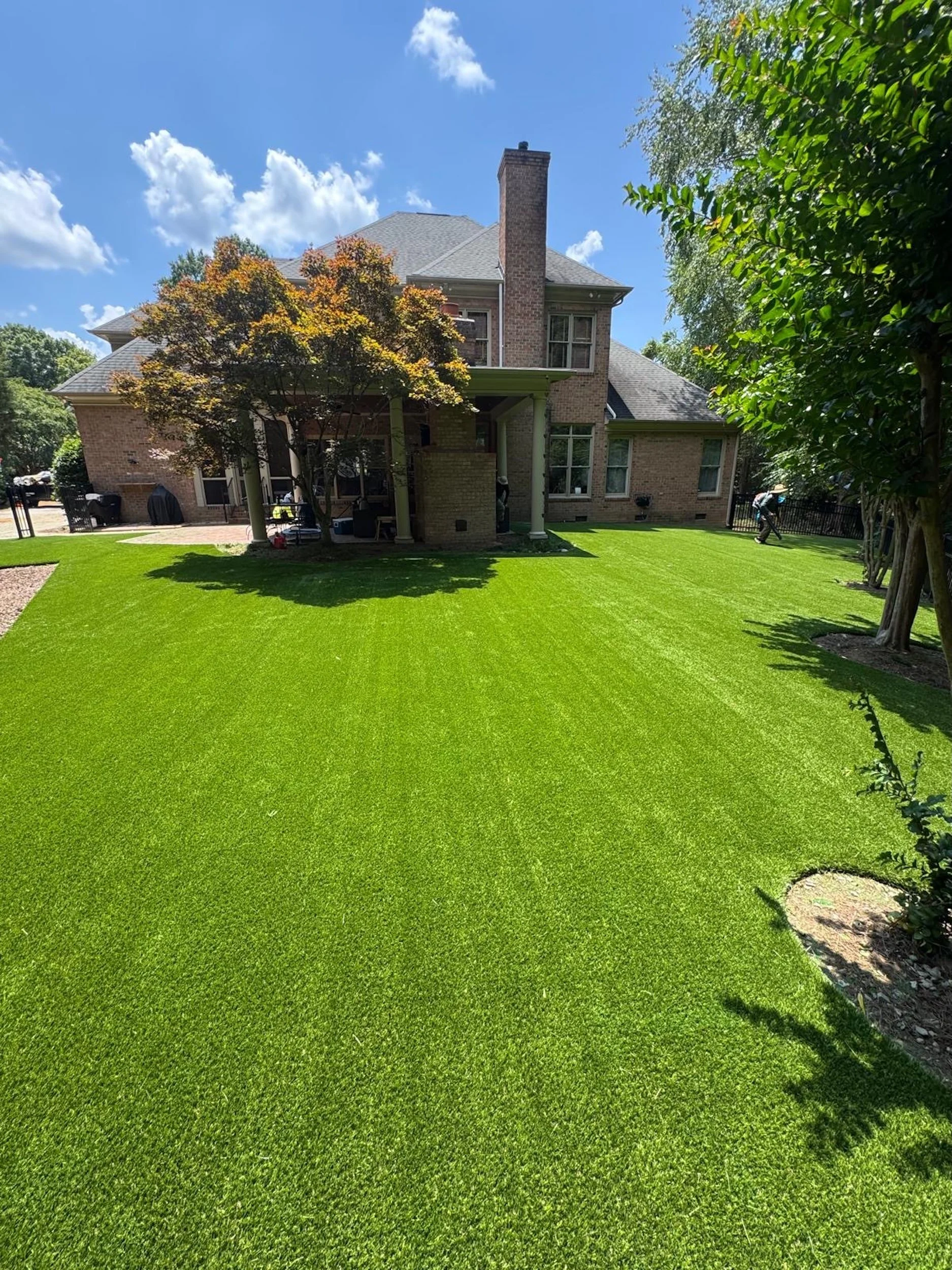 Backyard with meticulously maintained bright green grass, trees, a brick house, and a person working on yard in sunny weather.