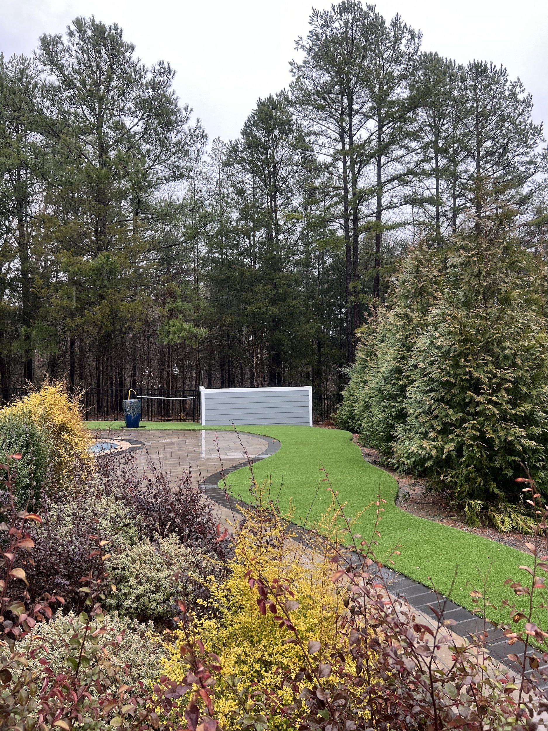 A backyard garden with a paved patio, decorative plants, a bright green artificial lawn, and tall trees in the background on an overcast day.