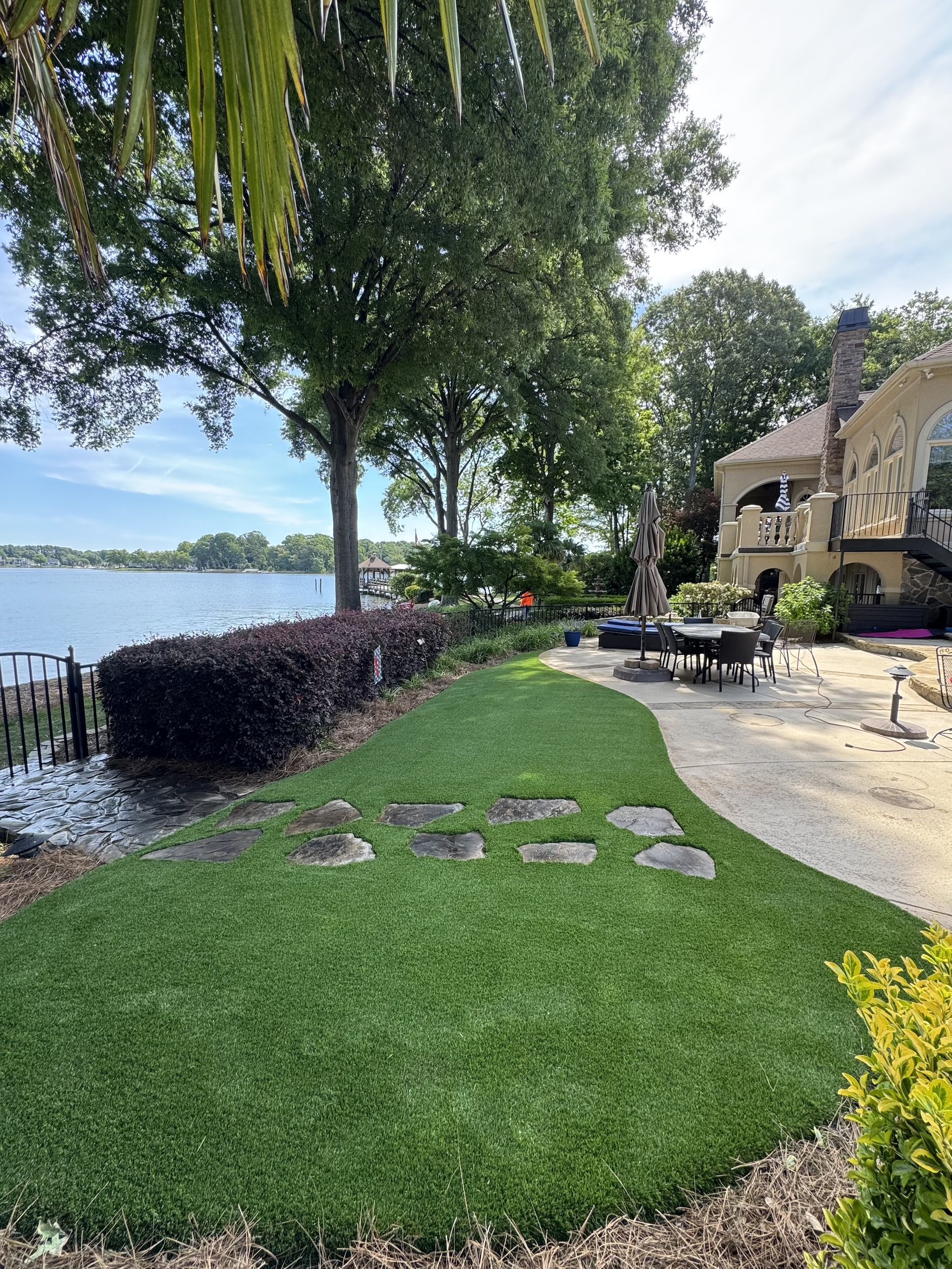 Lawn area with a stepping stone path, landscaped bushes, trees, and a house with a patio set overlooking a body of water with a dock in the distance.