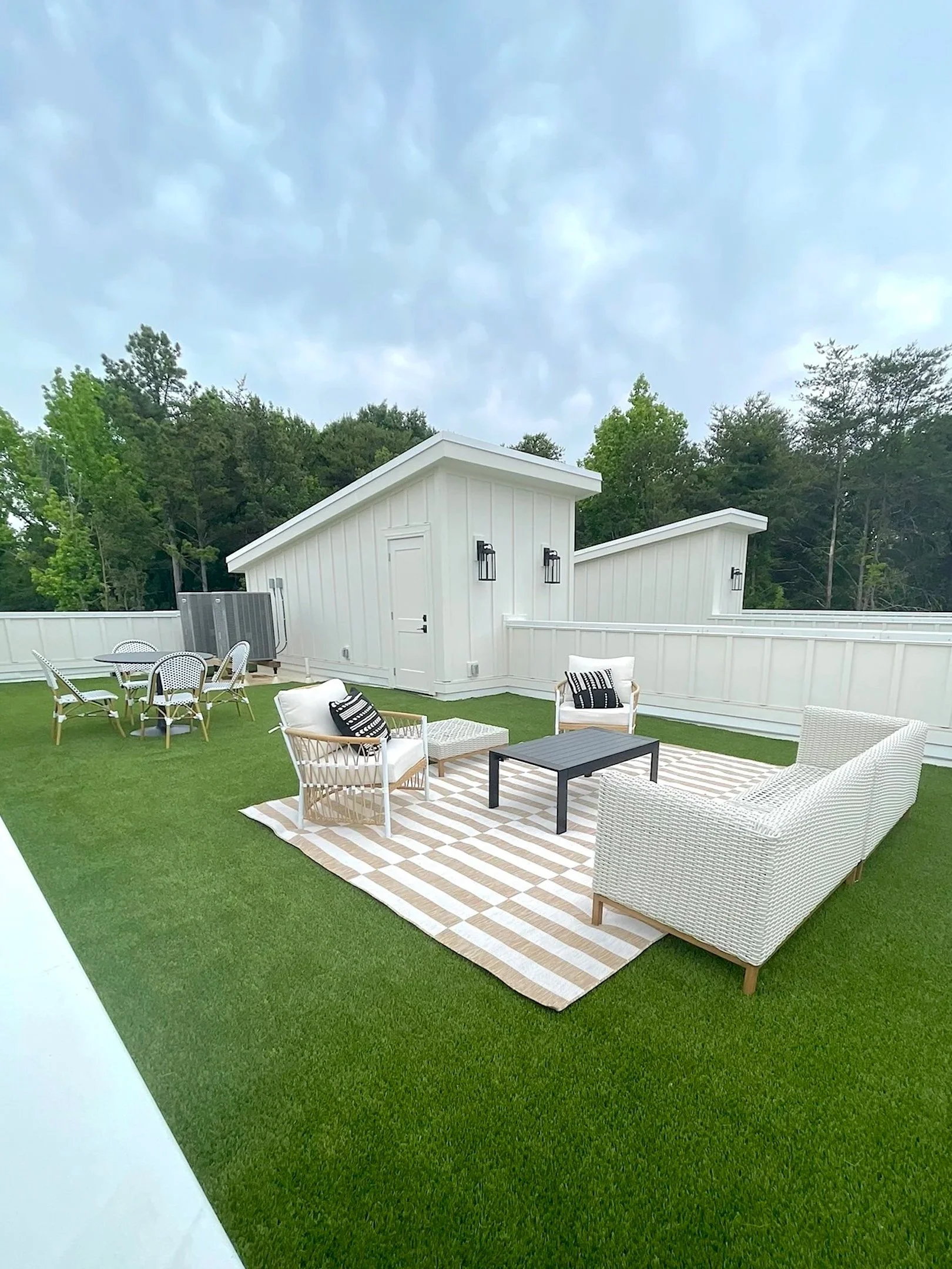 Backyard patio with outdoor furniture, including white patio chairs, a black table, a striped rug, a white shed, and a green lawn surrounded by a white fence and trees