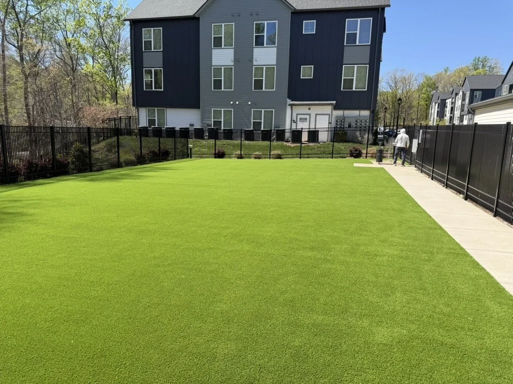 A well-maintained artificial grass lawn next to a concrete sidewalk, enclosed by a black metal fence. In the background, multi-story residential buildings and trees are visible under a clear blue sky.