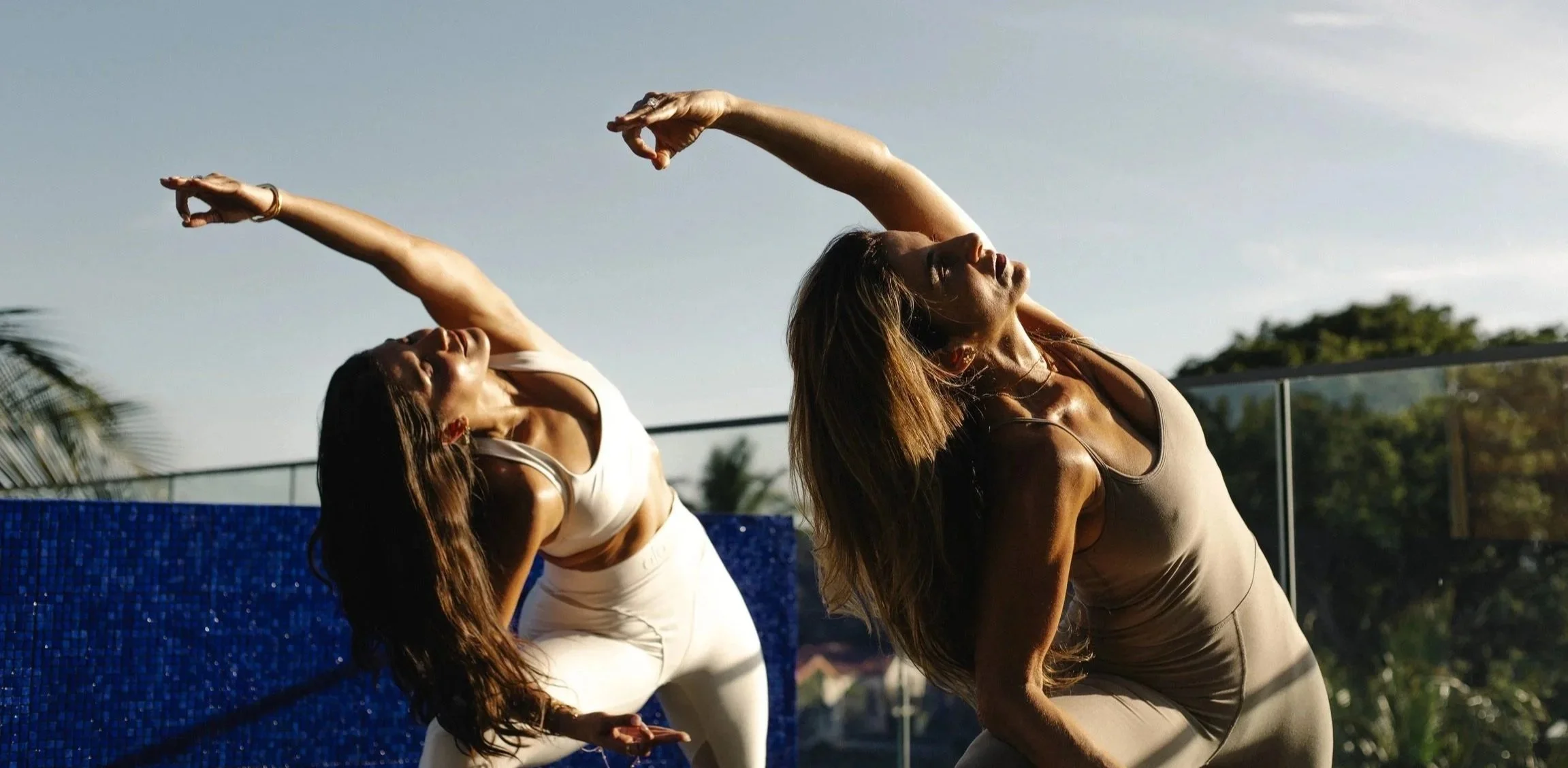 Two women practicing yoga outdoors during sunset, stretching and balancing on a rooftop with a blue mosaic wall behind them.