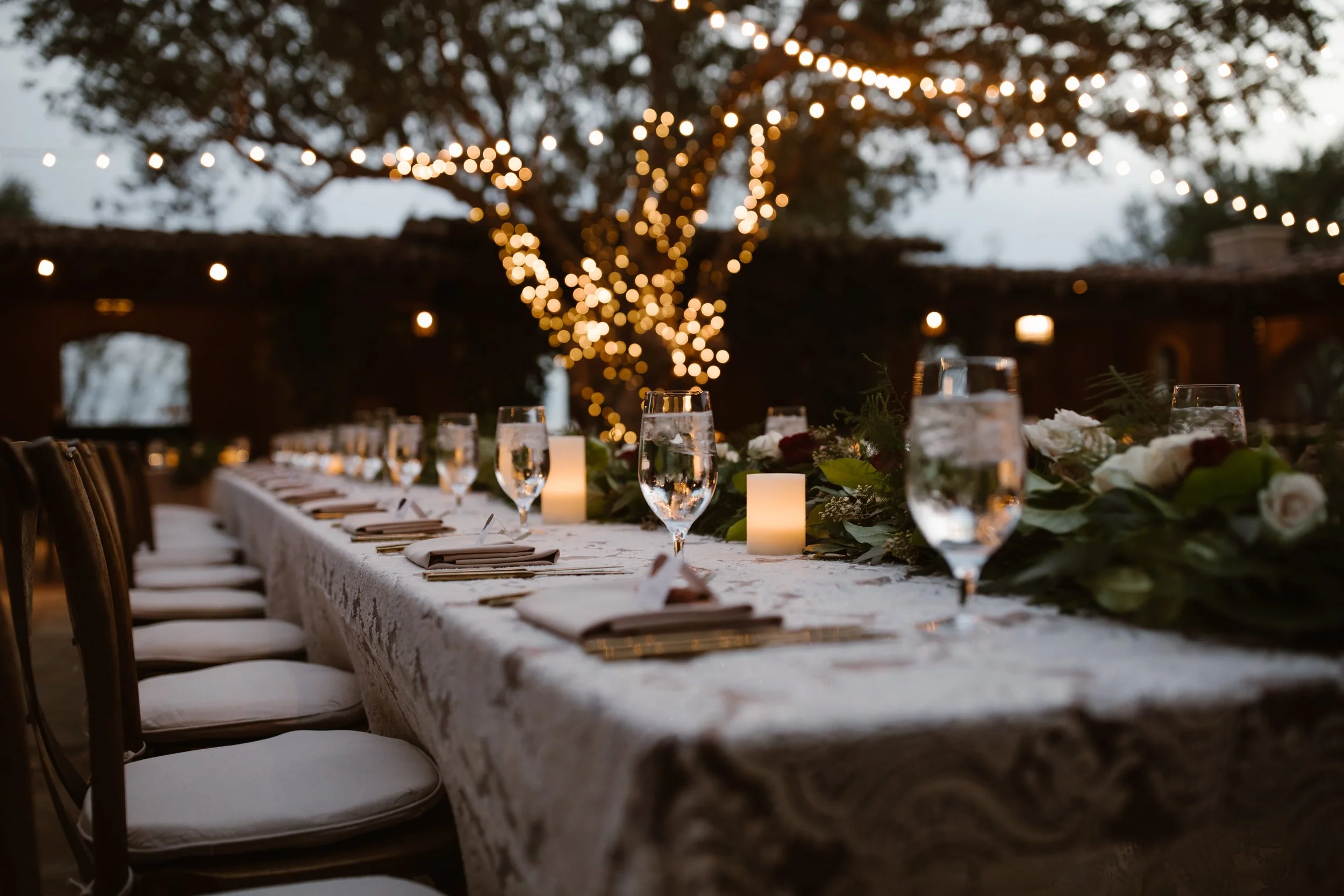 An outdoor dining table decorated with glasses, candles, and floral arrangements, set for an evening meal under string lights on a tree.