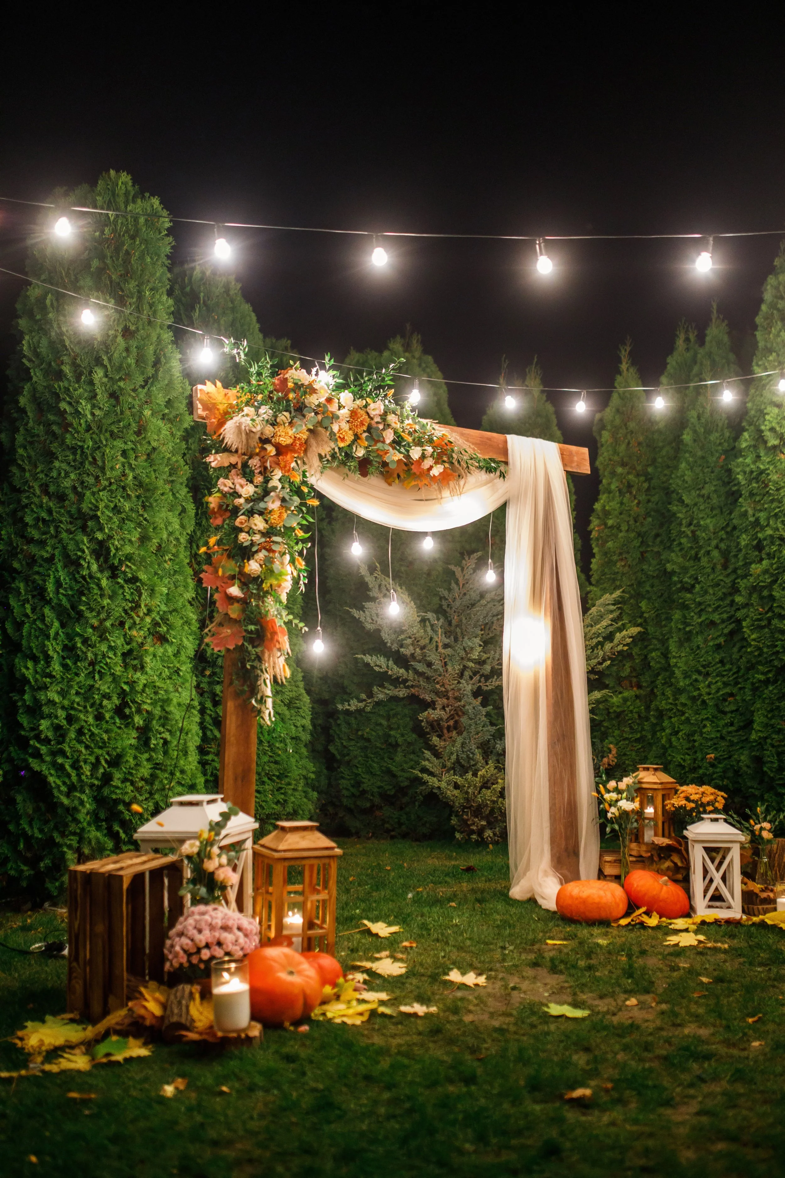 Night outdoor wedding setup with an rustic wooden arch decorated with flowers and draped fabric, string lights overhead, surrounded by tall green trees, pumpkins, lanterns, candles, and autumn leaves.