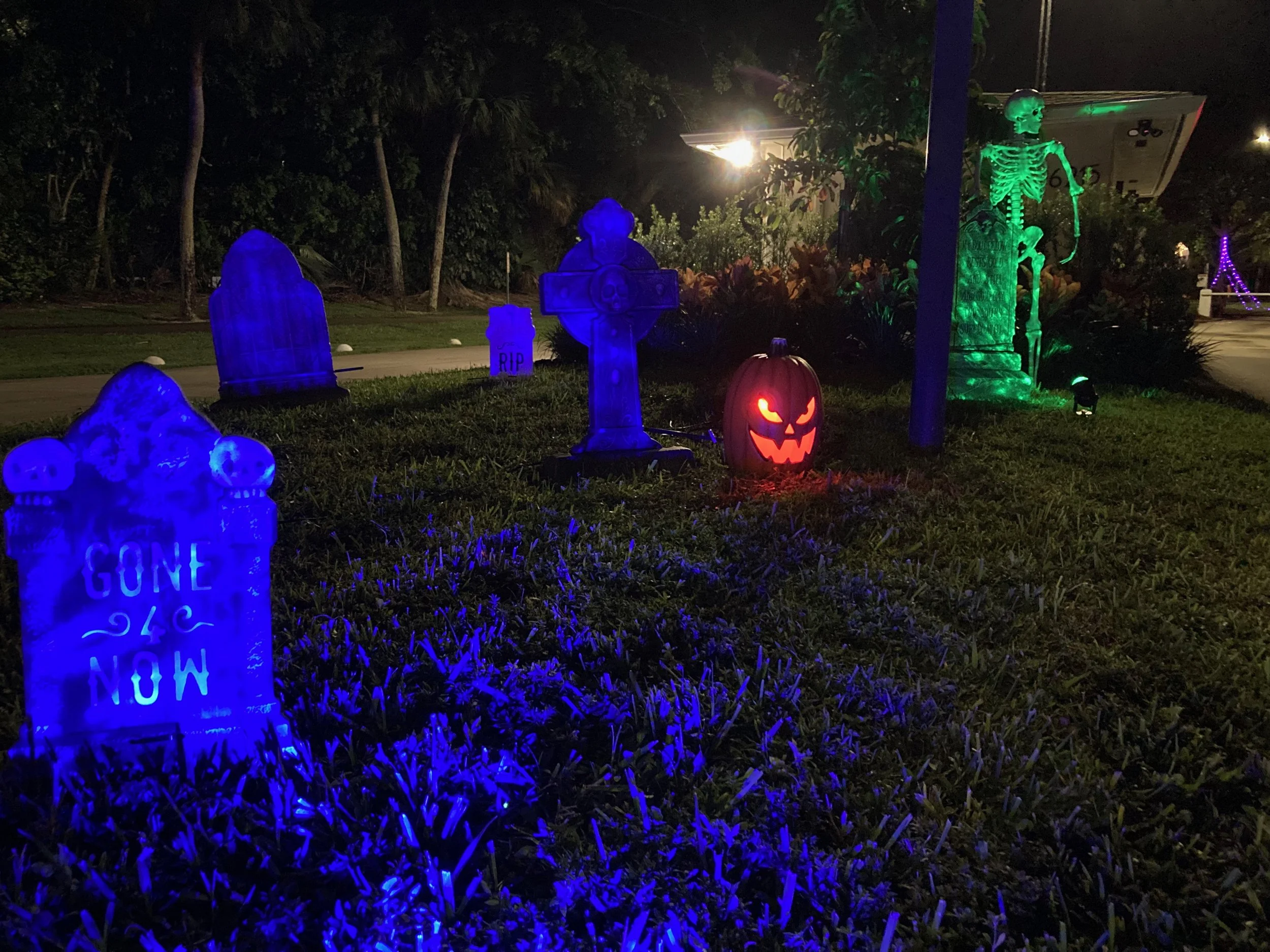 Halloween yard decorations at night, including illuminated tombstones, a decorated pumpkin with a scary face, a skeleton illuminated in green, and other spooky decorations.
