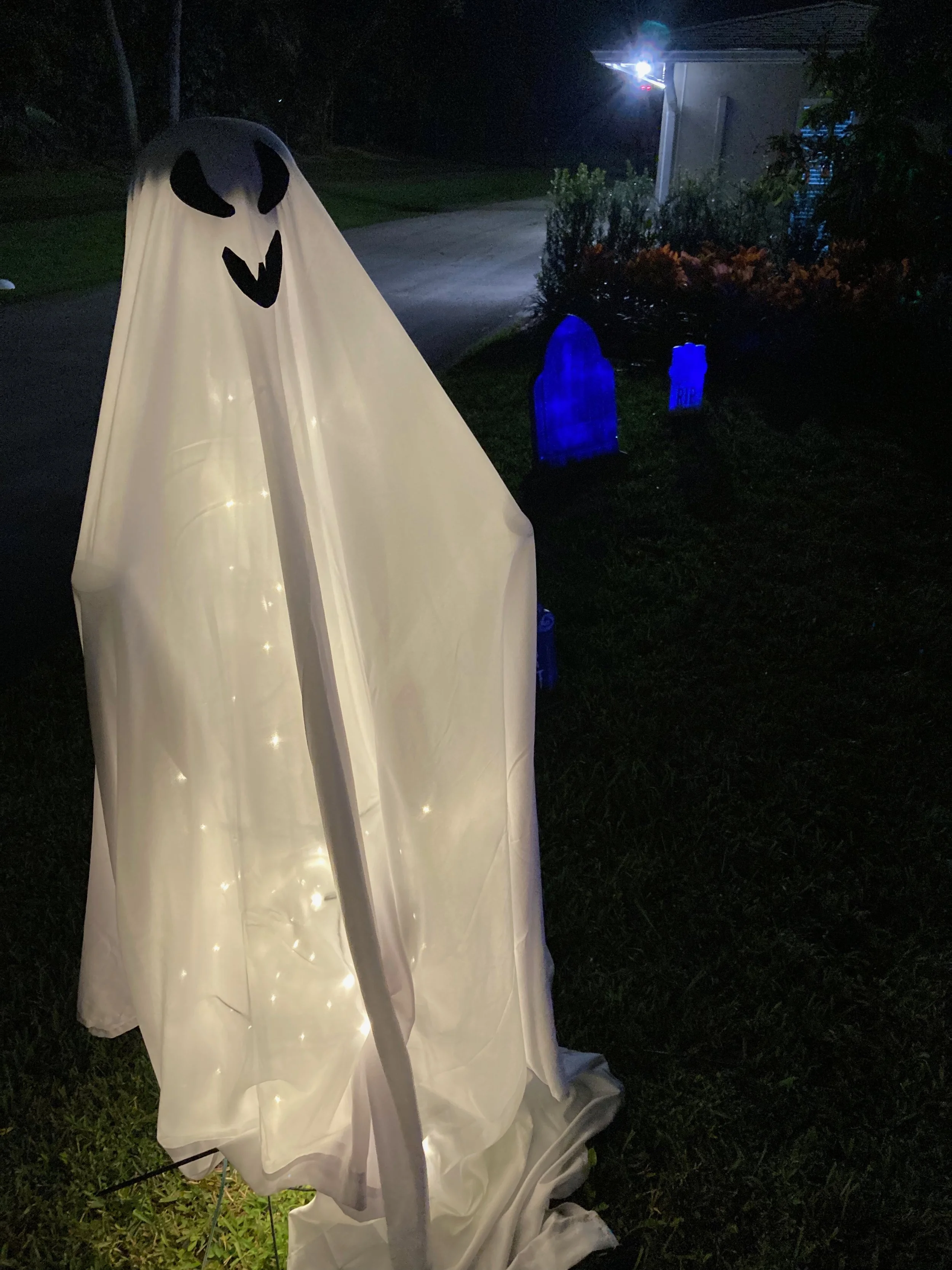 A white sheet with a ghost face drawn on it, illuminated from below, positioned outdoors at night near some grave markers and bushes.