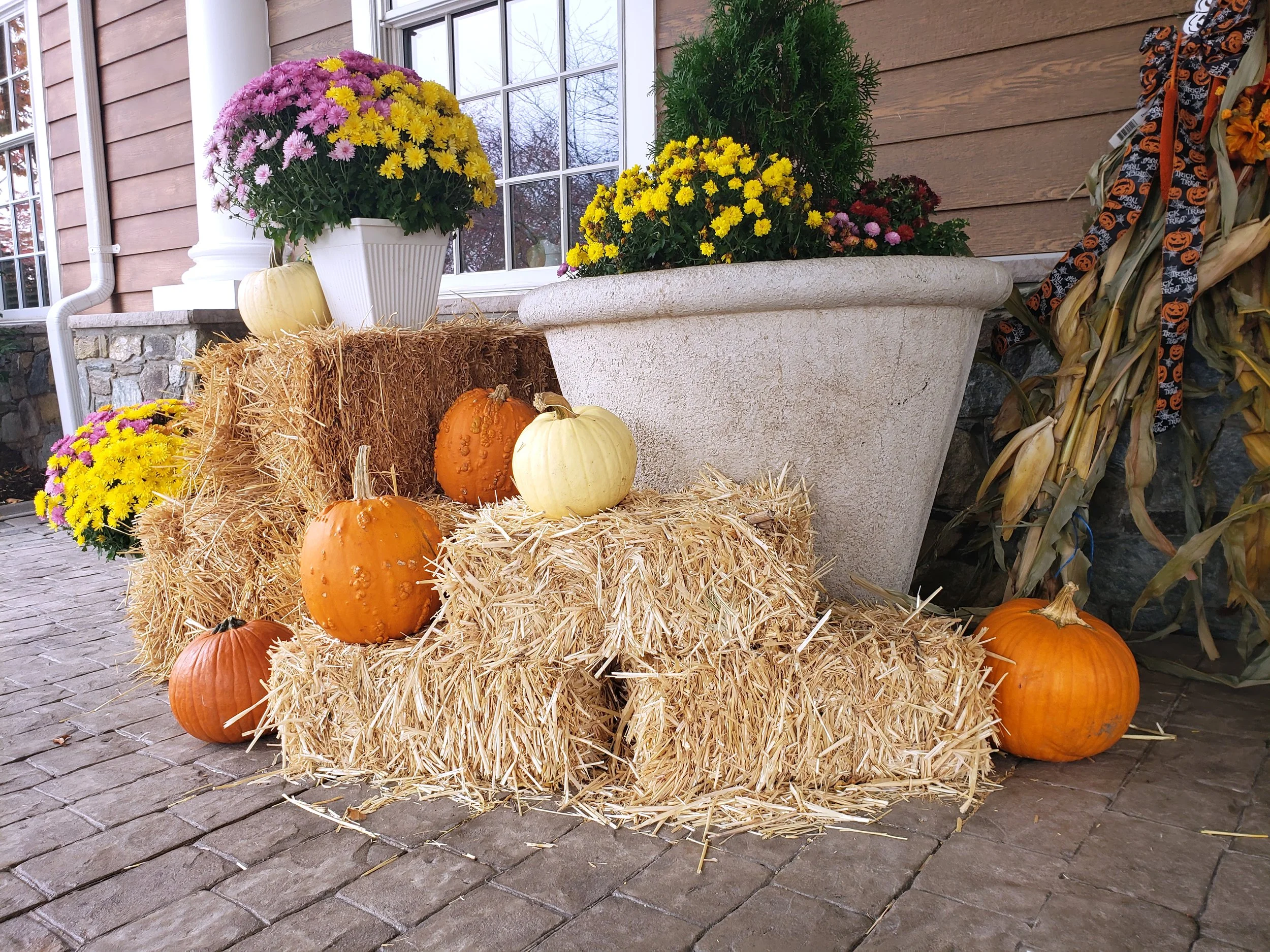 Fall-themed display with pumpkins, hay bales, and flower pots with yellow and pink flowers on a porch with a stone and wooden exterior.