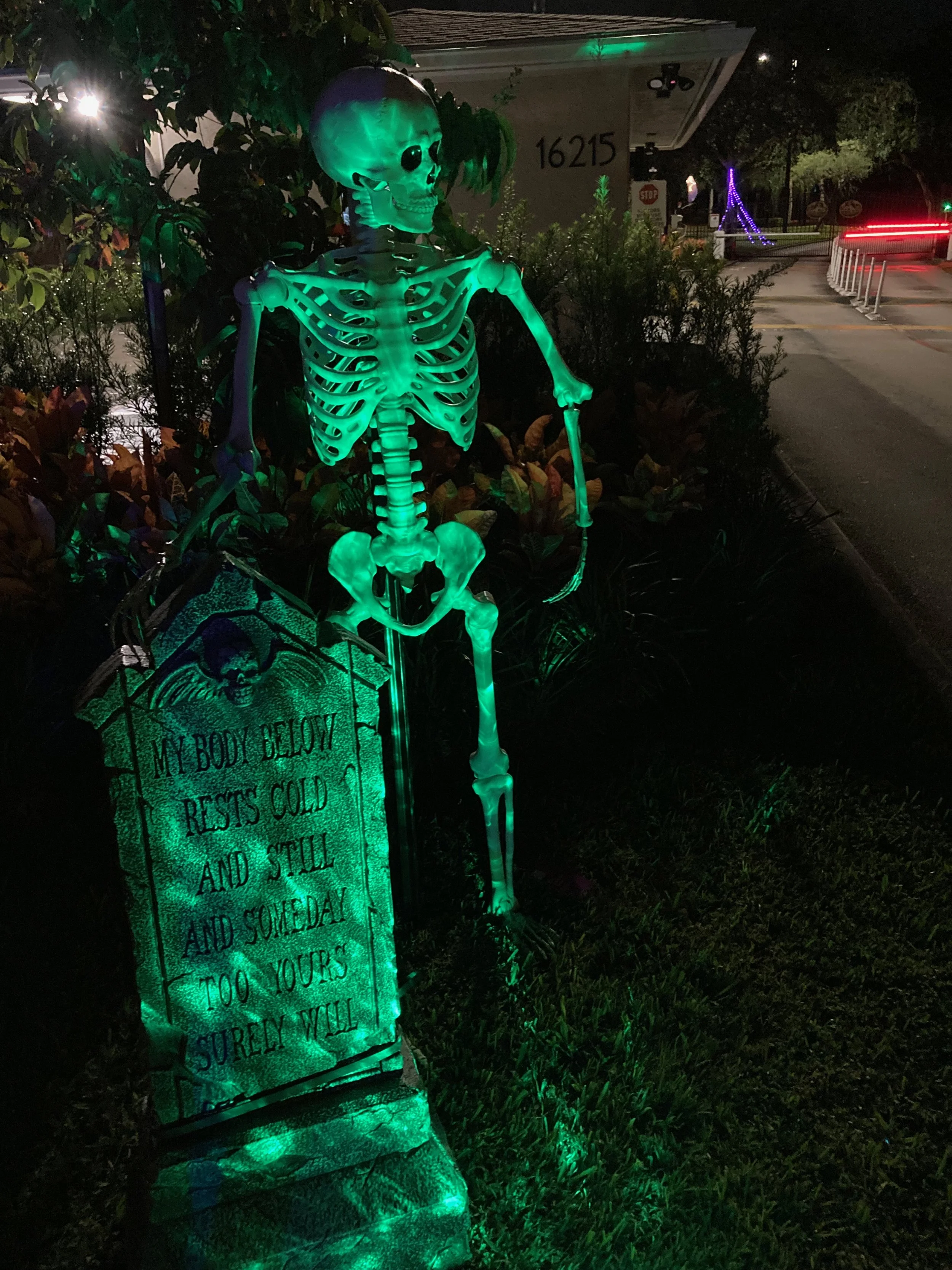 A glowing green skeleton decoration standing outdoors at night, illuminated from below, with a tombstone that reads, 'My body below rests cold and still, tomorrow and someday, but your love will surely will.'