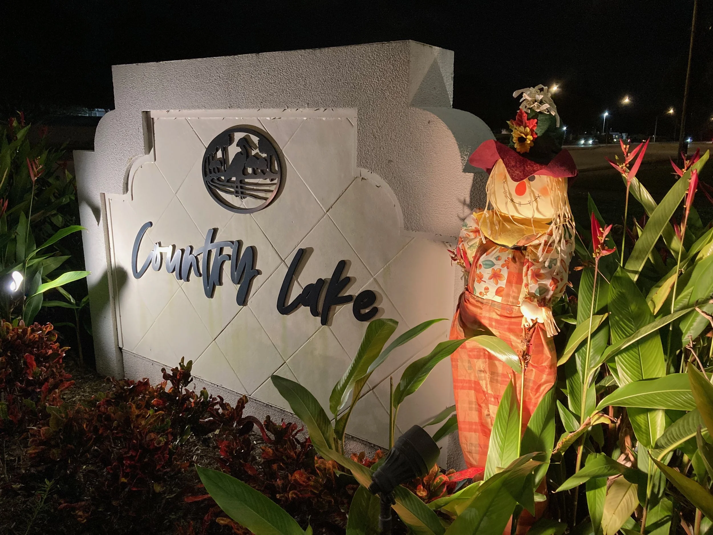Nighttime scene featuring a white concrete structure with a circular cutout displaying a tree-like design and the words 'Country Lake' in script. A scarecrow decoration with a floral head, black hat, red cape, and orange checkered pants is visible, surrounded by green plants and red foliage, with streetlights in the background.