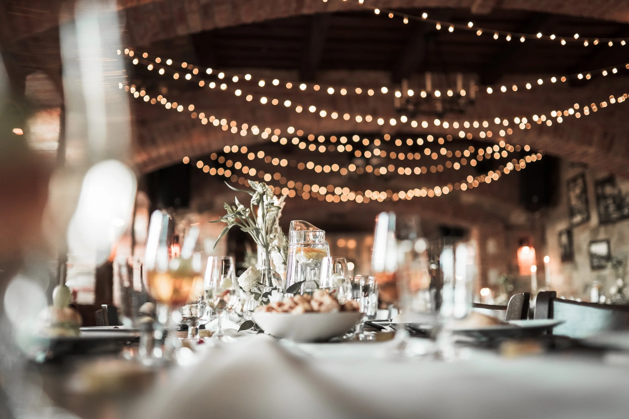 Wedding reception in a rustic venue with string lights hanging from the wooden ceiling, decorated tables with floral centerpieces, glassware, and table settings.