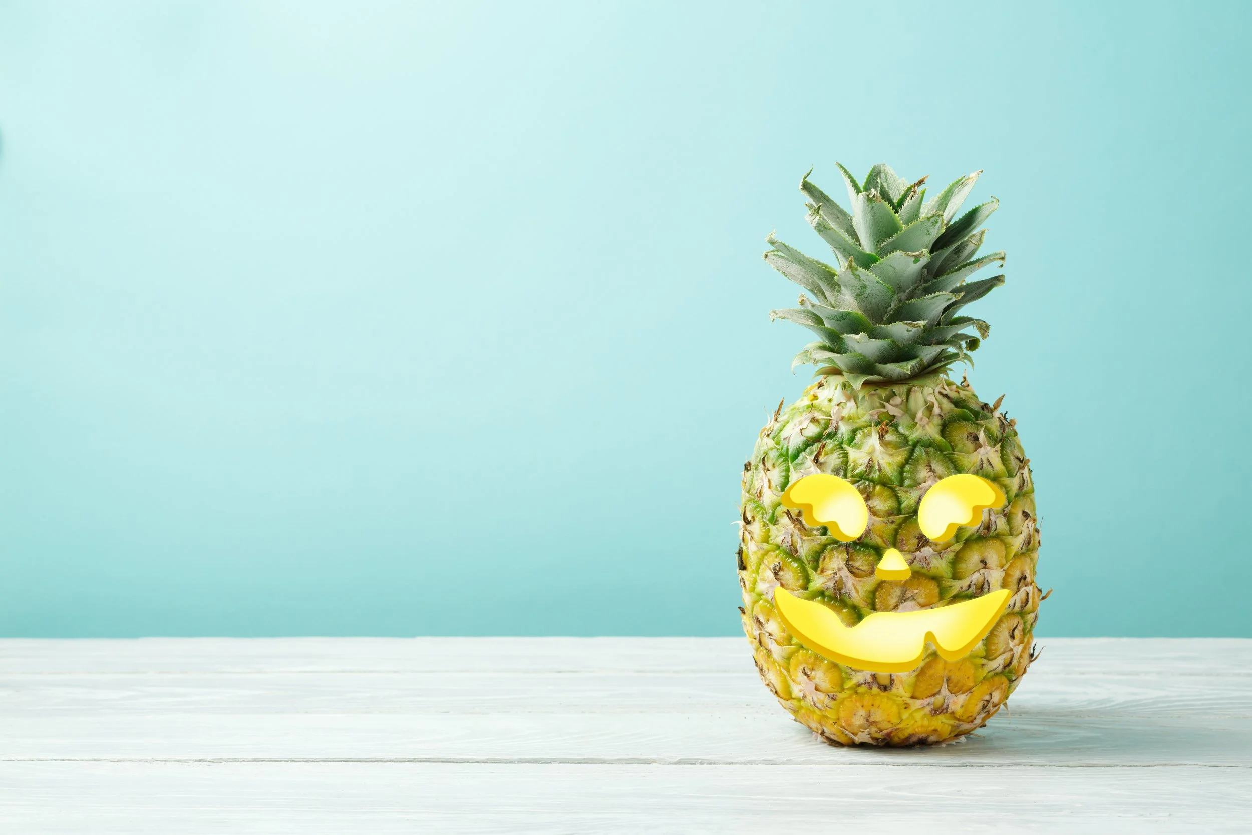 A pineapple with a smiley face carved into it, placed on a white wooden surface against a light blue background.