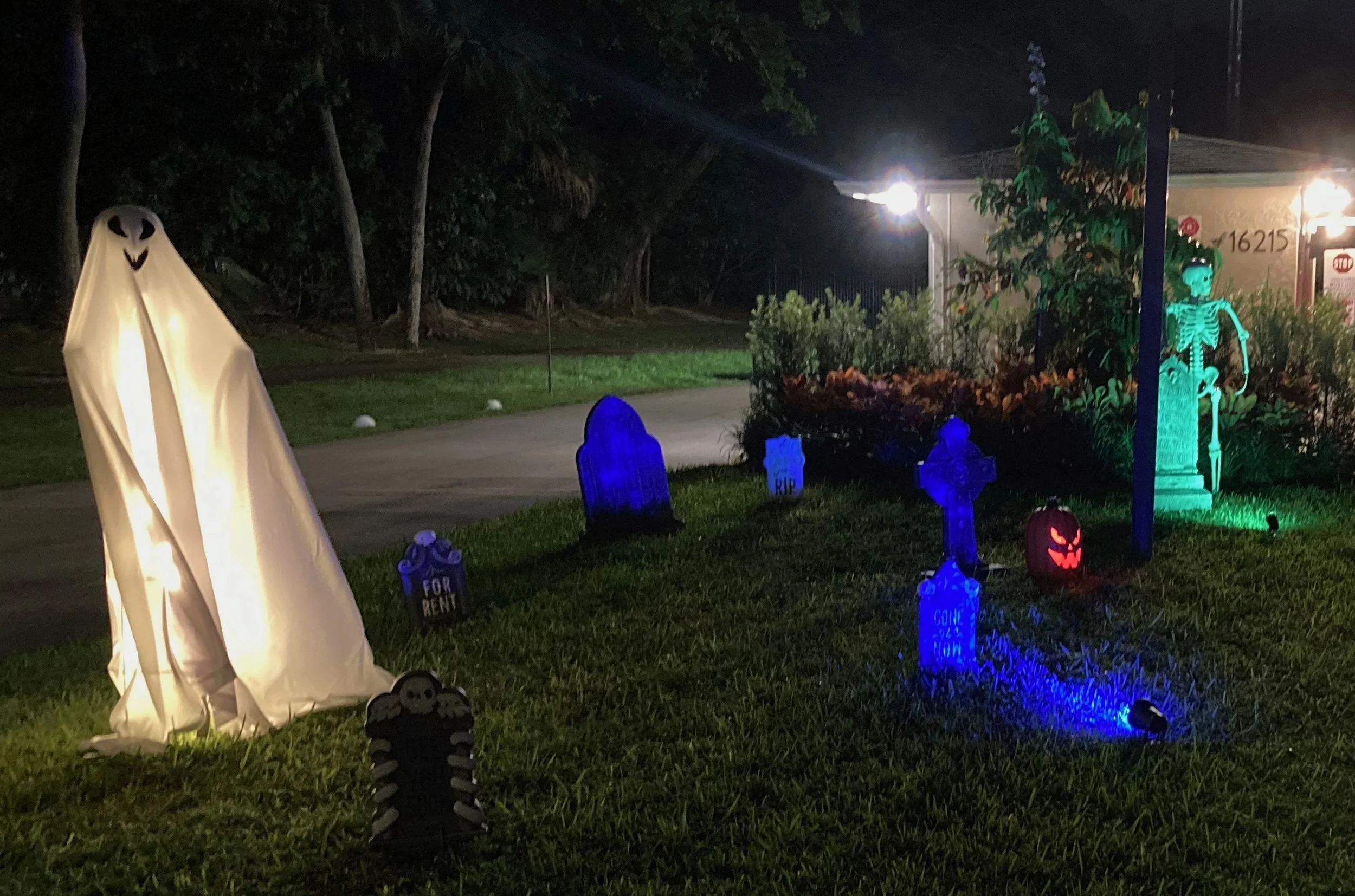 Nighttime Halloween yard decoration with ghost, skeletons, skulls, pumpkins, and glow-in-the-dark signs.