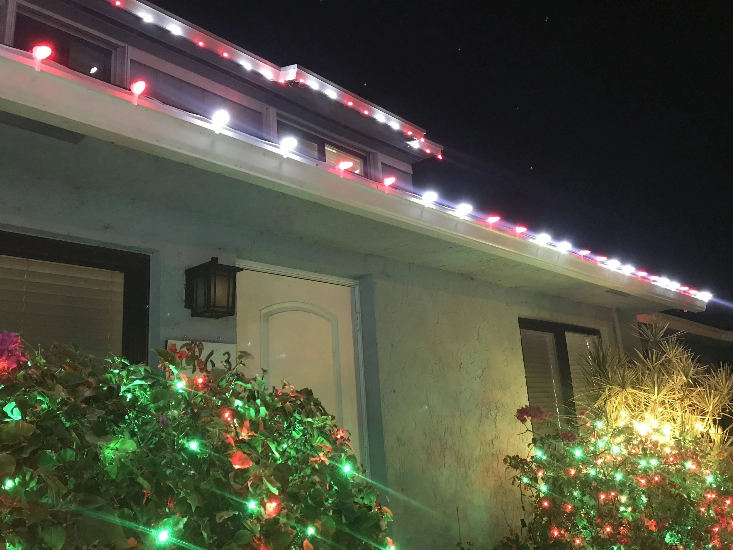 Nighttime exterior of a house decorated with red and white Christmas string lights along the roofline, with bushes decorated with red and green Christmas lights in the foreground.