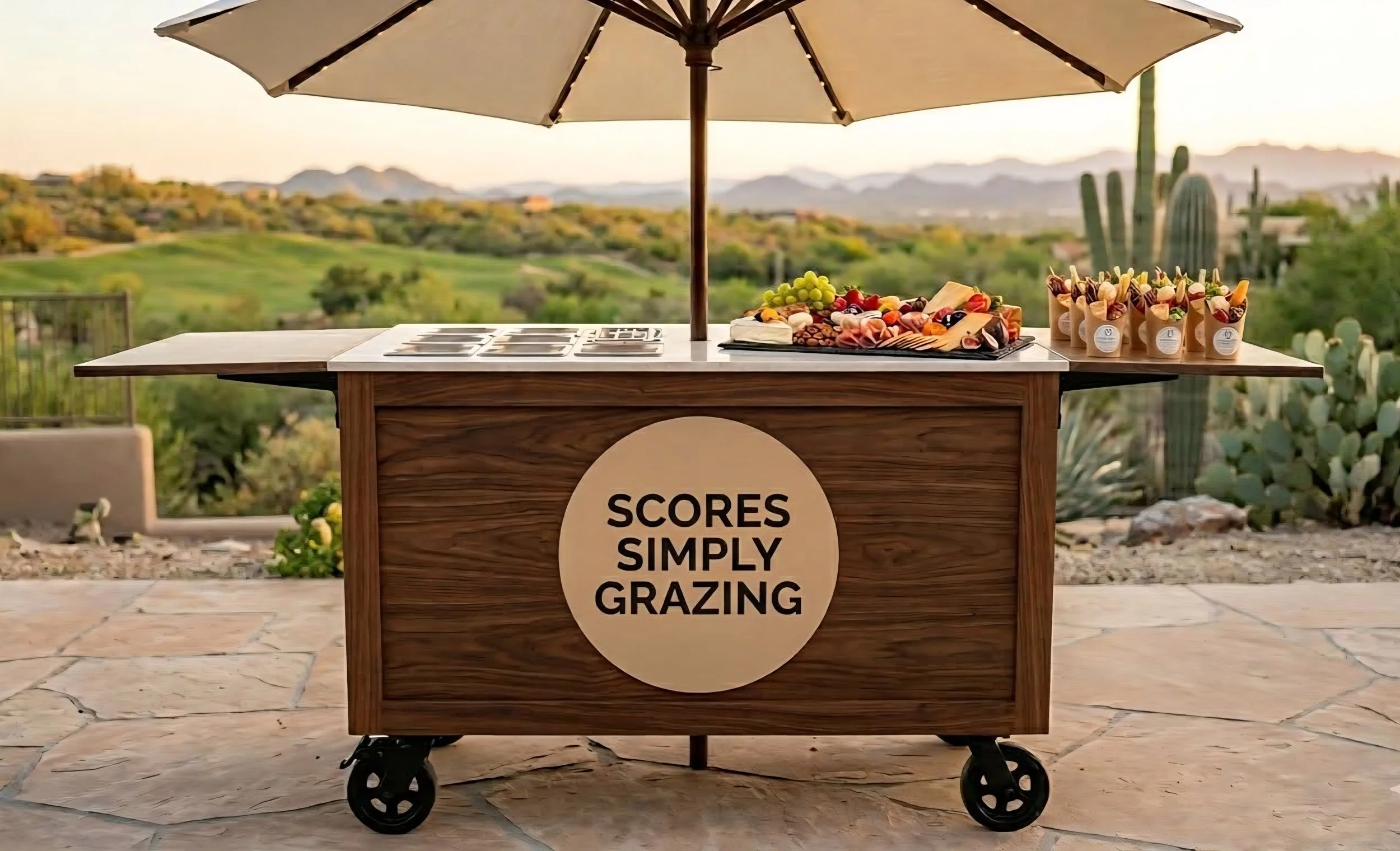 A wooden cart with a large round sign that reads 'SCORES SIMPLY GRAZING'. The cart is set outdoors on a stone patio with a scenic background of greenery, mountains, and cacti under a large patio umbrella. The cart is topped with grape and cheese arrangements and small containers of colorful snacks.