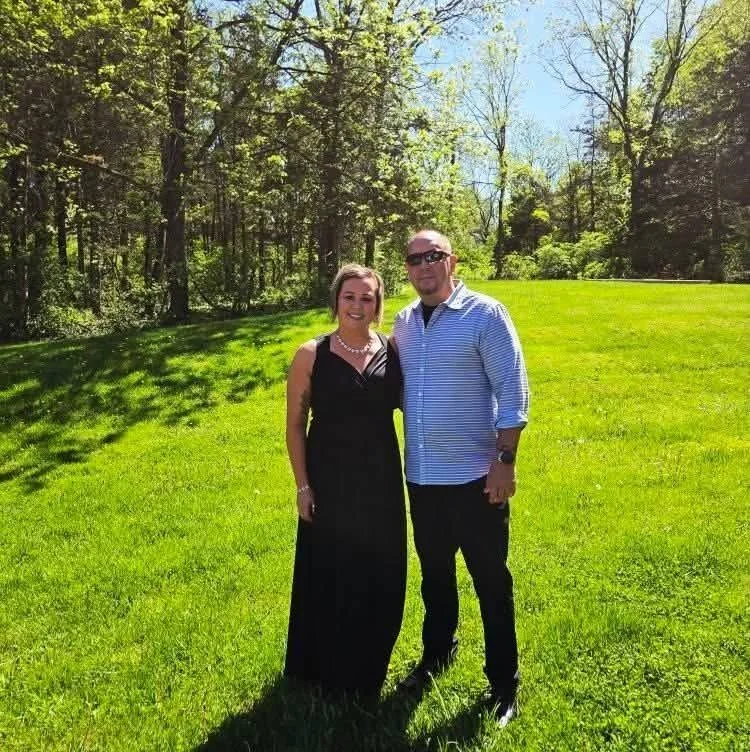 A woman in a black dress and a man in a blue striped shirt pose together outdoors on a bright sunny day, standing on green grass with trees in the background.