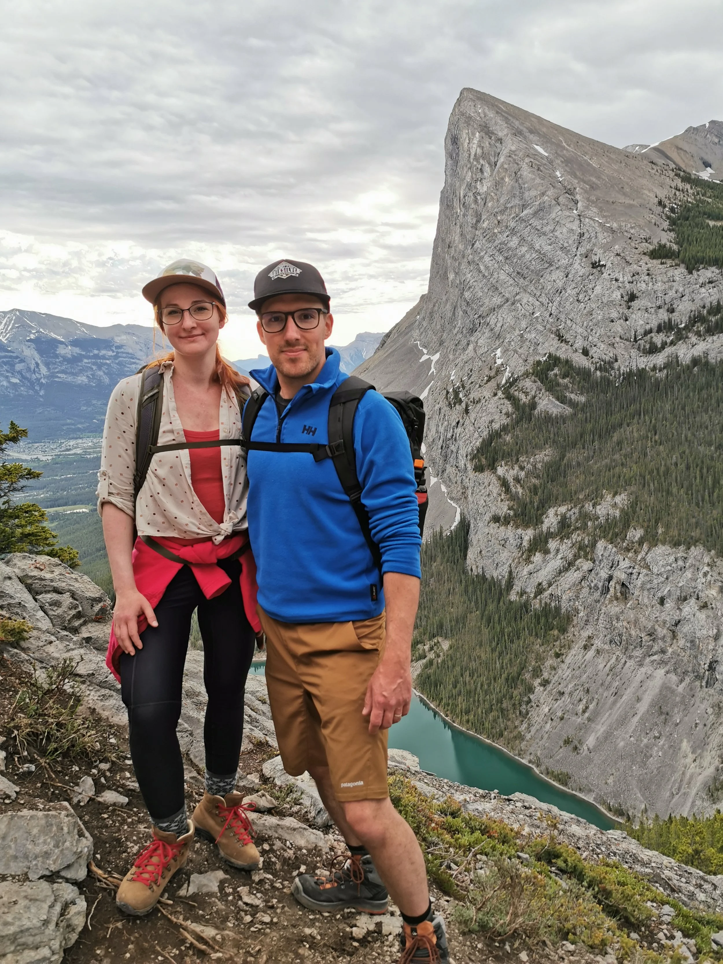 A man and woman hiking on a rocky trail in the mountains with a large cliff, forest, and lake in the background. Both are wearing outdoor clothing and backpacks.