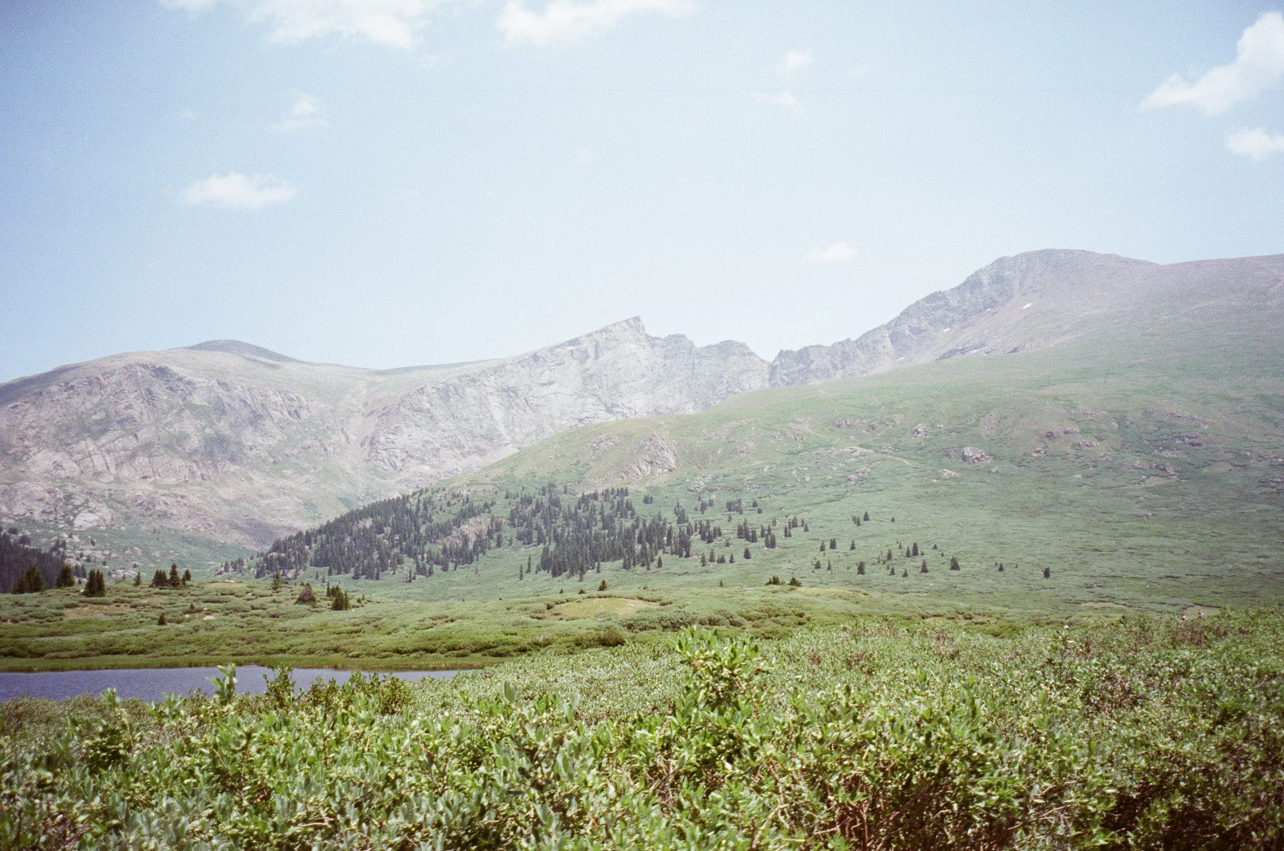 A landscape photo of green mountains with rocky peaks, a body of water in the foreground, and a blue sky with scattered clouds.