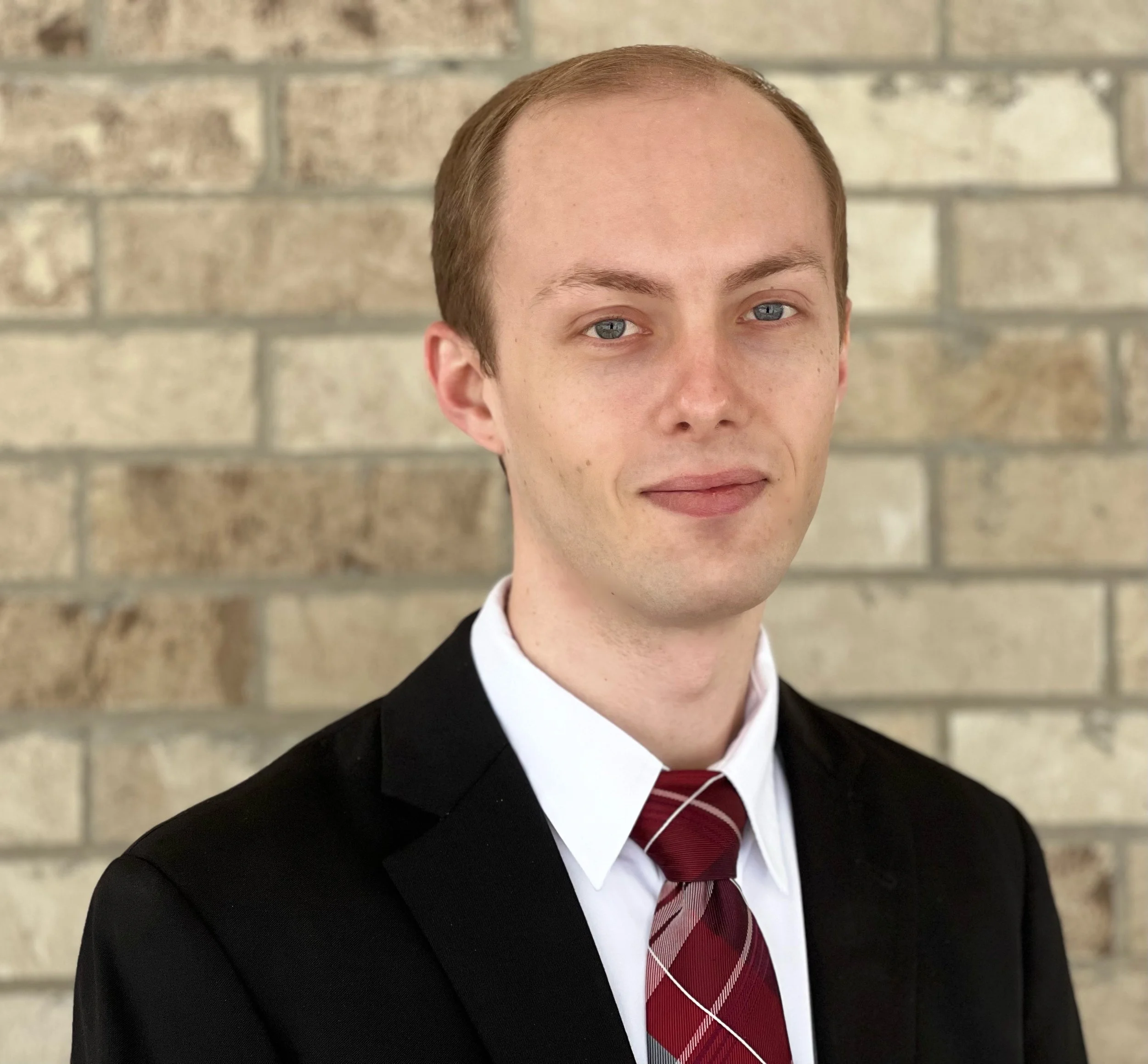 A young man in formal attire, with a black suit, white shirt, and red tie, standing against a brick wall background.