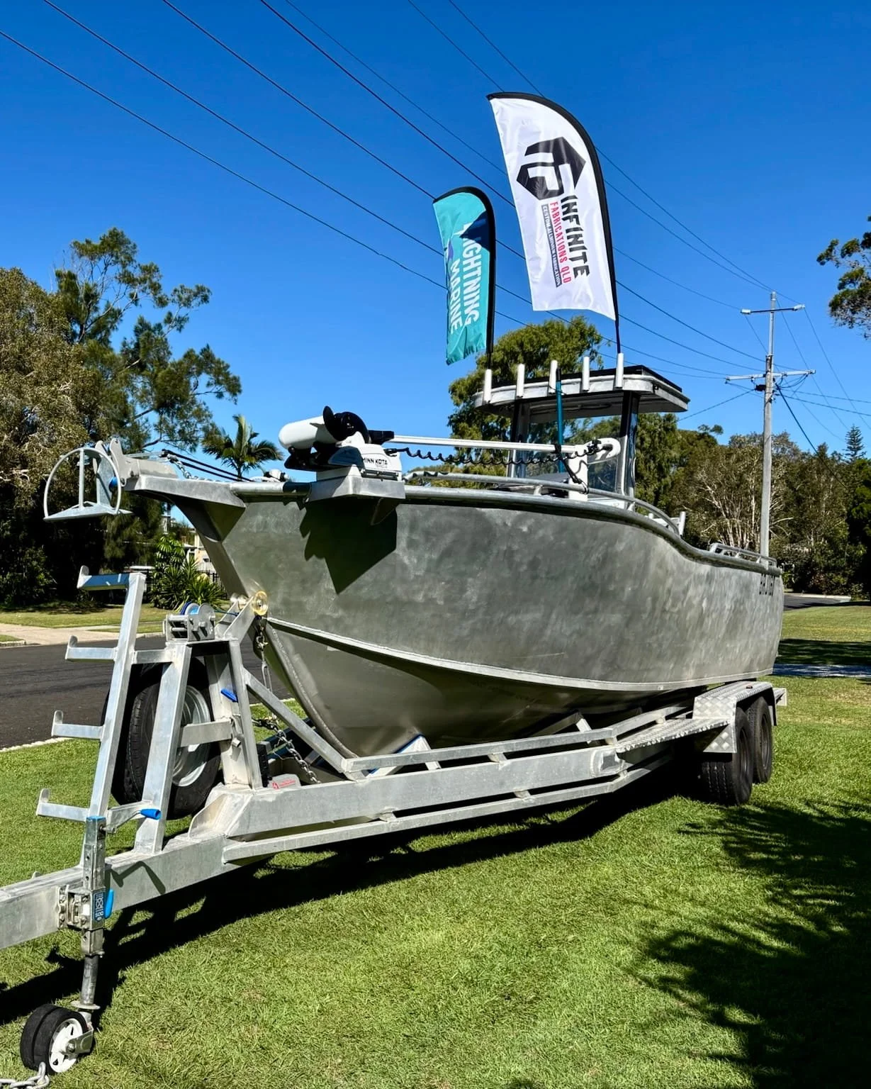 A silver boat on a trailer parked on a grassy area with trees and utility poles in the background. There are flags on the boat, one with a logo and the other with the text 'Lightfish Fiberglass, LLC'.